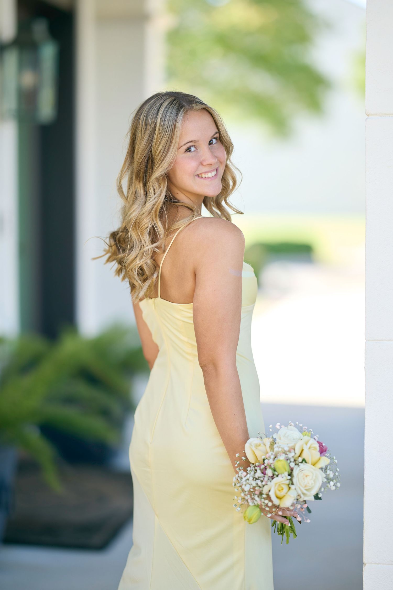 Young woman in yellow dress holding bouquet of flowers, smiling over shoulder outdoors.
