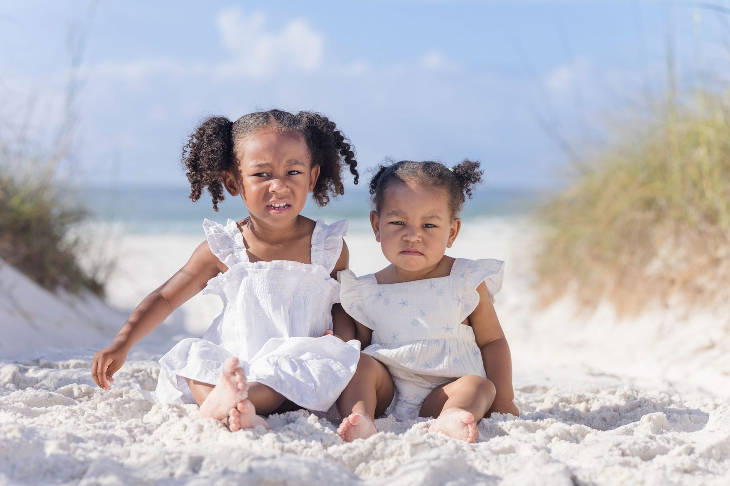 Two young girls in white dresses sitting together on white sand beach, smiling happily on a sunny day.