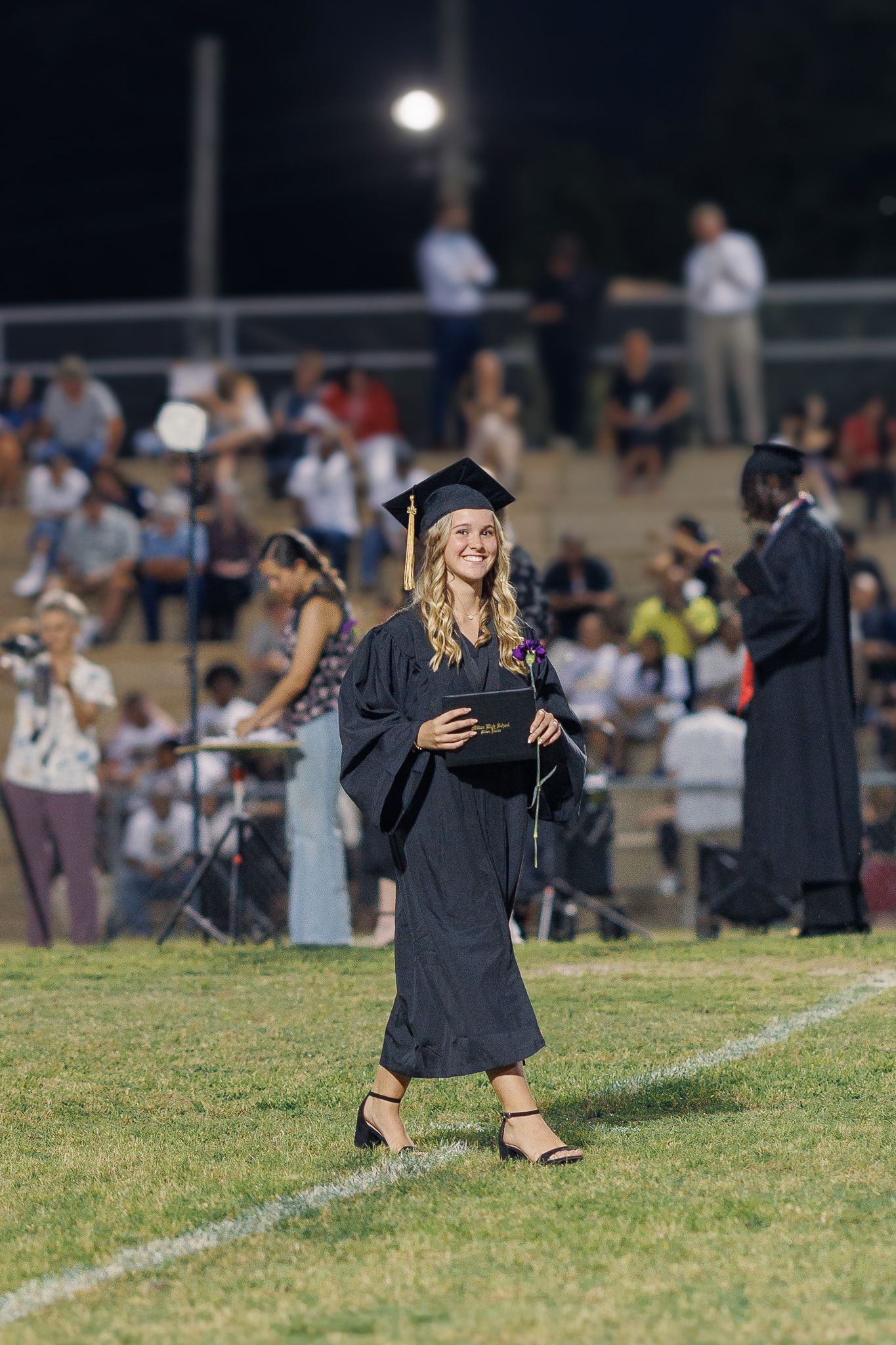 Graduate poses on field at night during outdoor commencement ceremony with crowd in background.