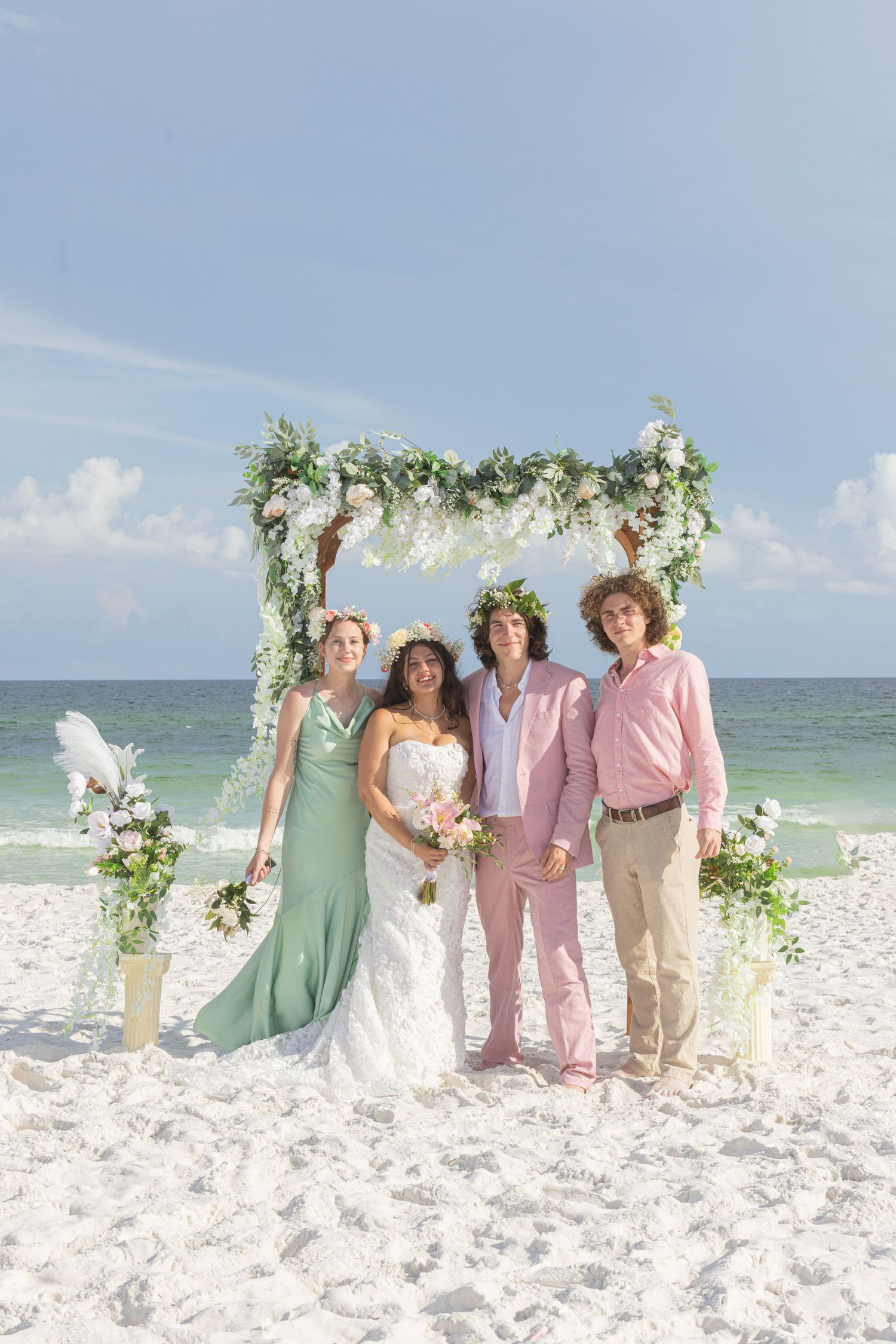 Wedding party poses under floral arch on beach with mint green and pink color scheme.