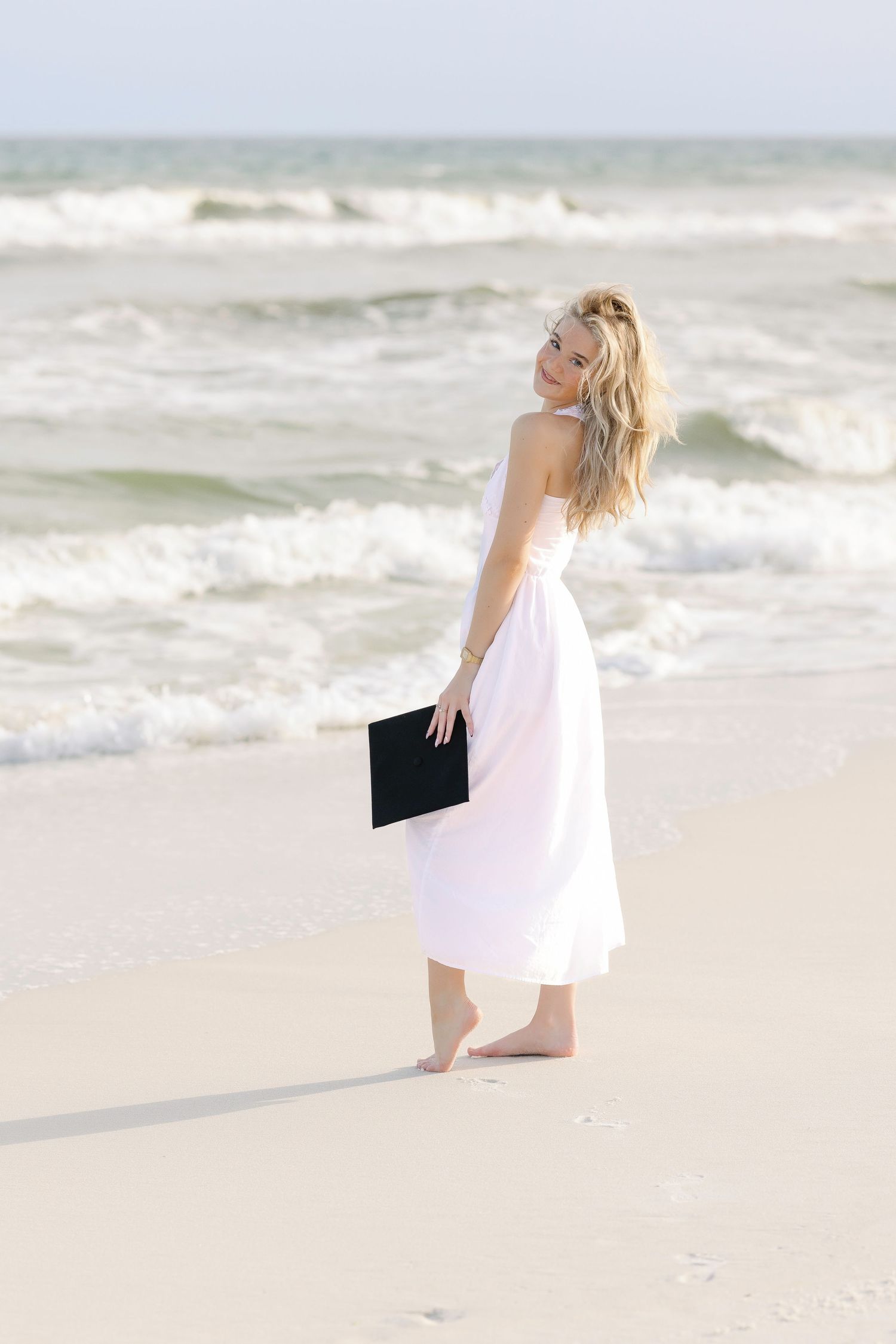 Person in flowing white dress standing on beach holding black folder while facing ocean waves.