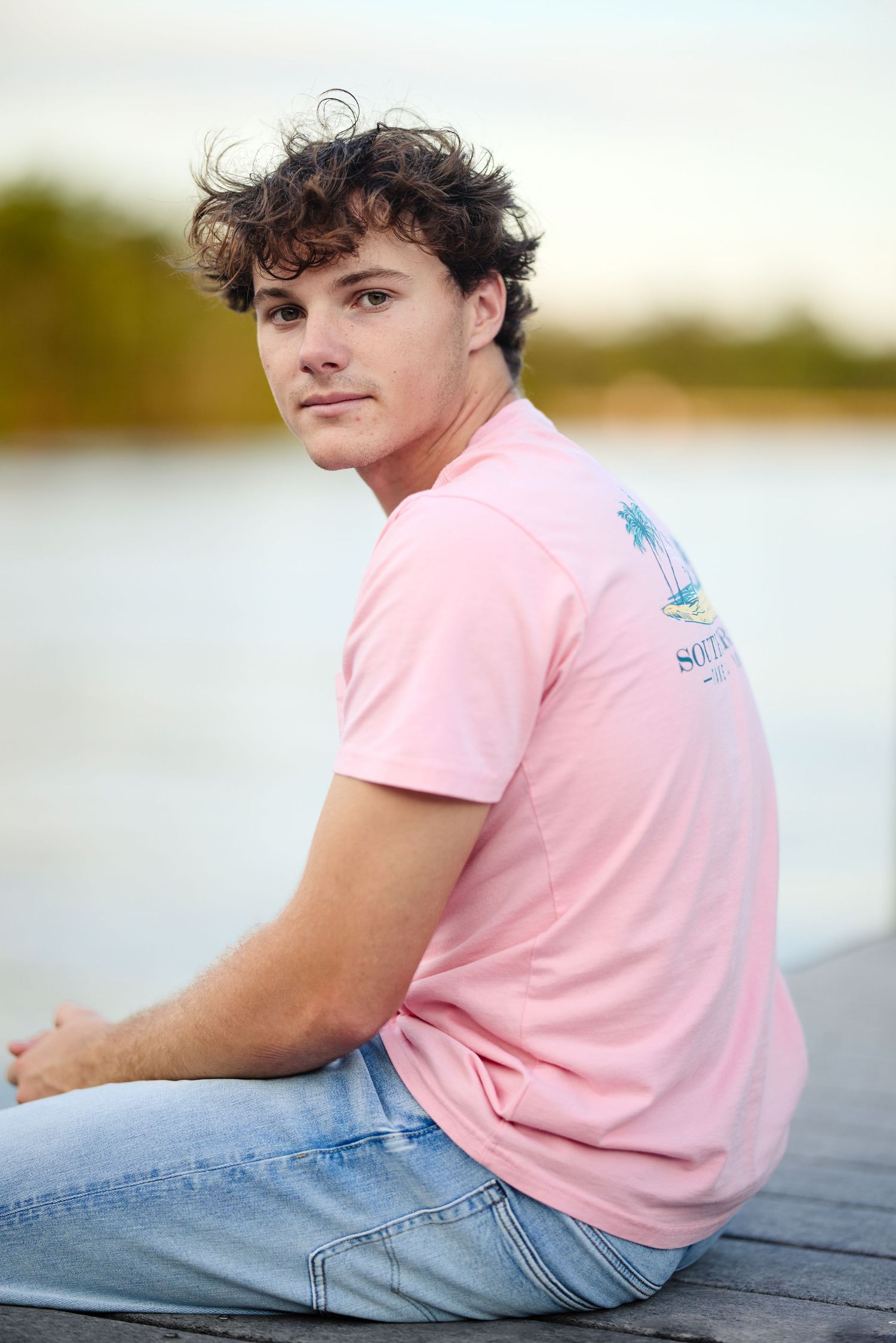 Person in pink t-shirt sitting on dock with water and trees in background.