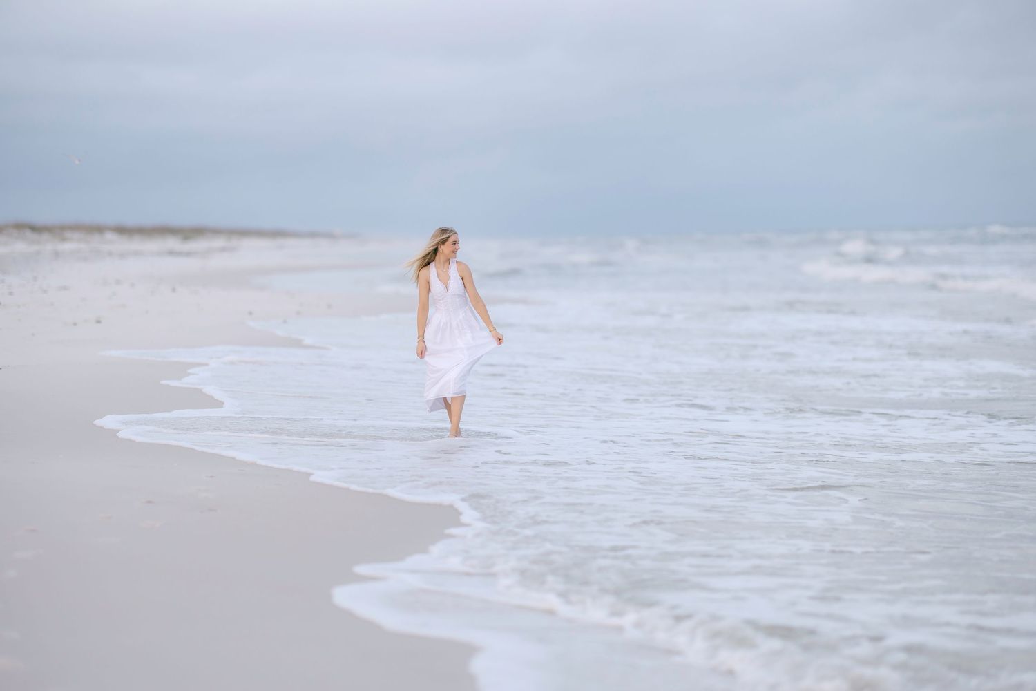 Figure in white dress walks along the shoreline on a misty beach.