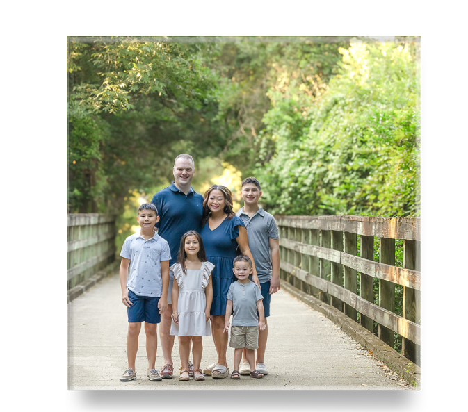 Family of six posing on a wooden bridge path surrounded by lush green trees in an outdoor portrait.