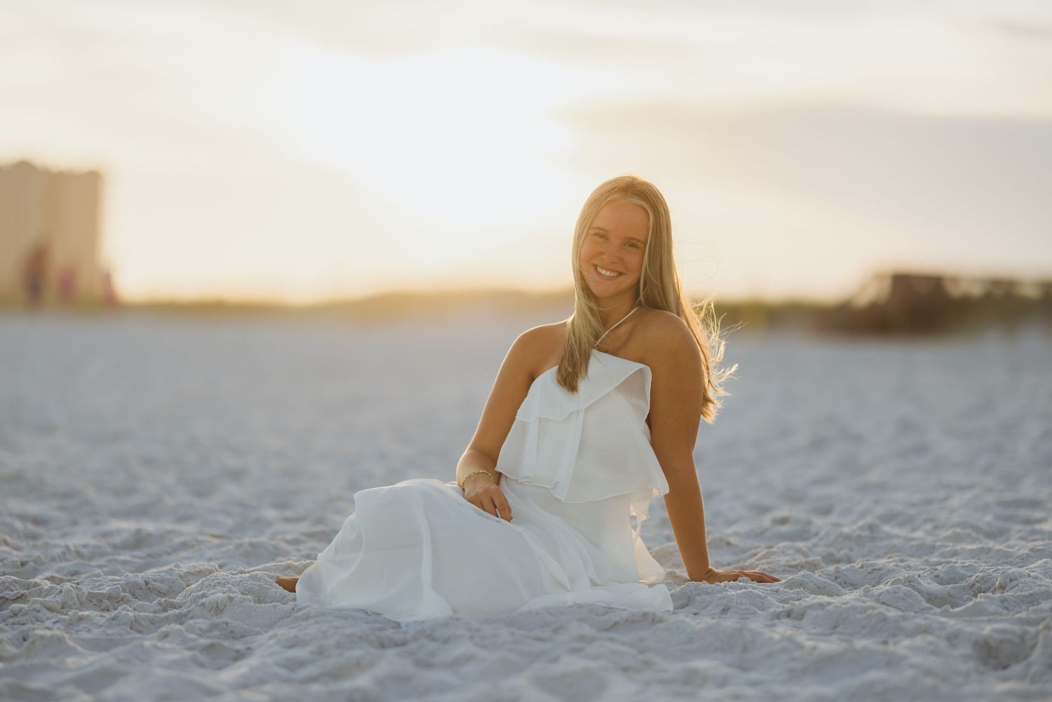 Smiling young woman in white dress sitting on sandy beach at golden sunset.