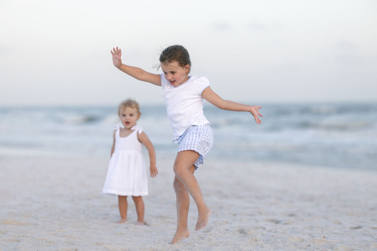 Two young girls playing joyfully on a sandy beach, one jumping with arms raised near the ocean.