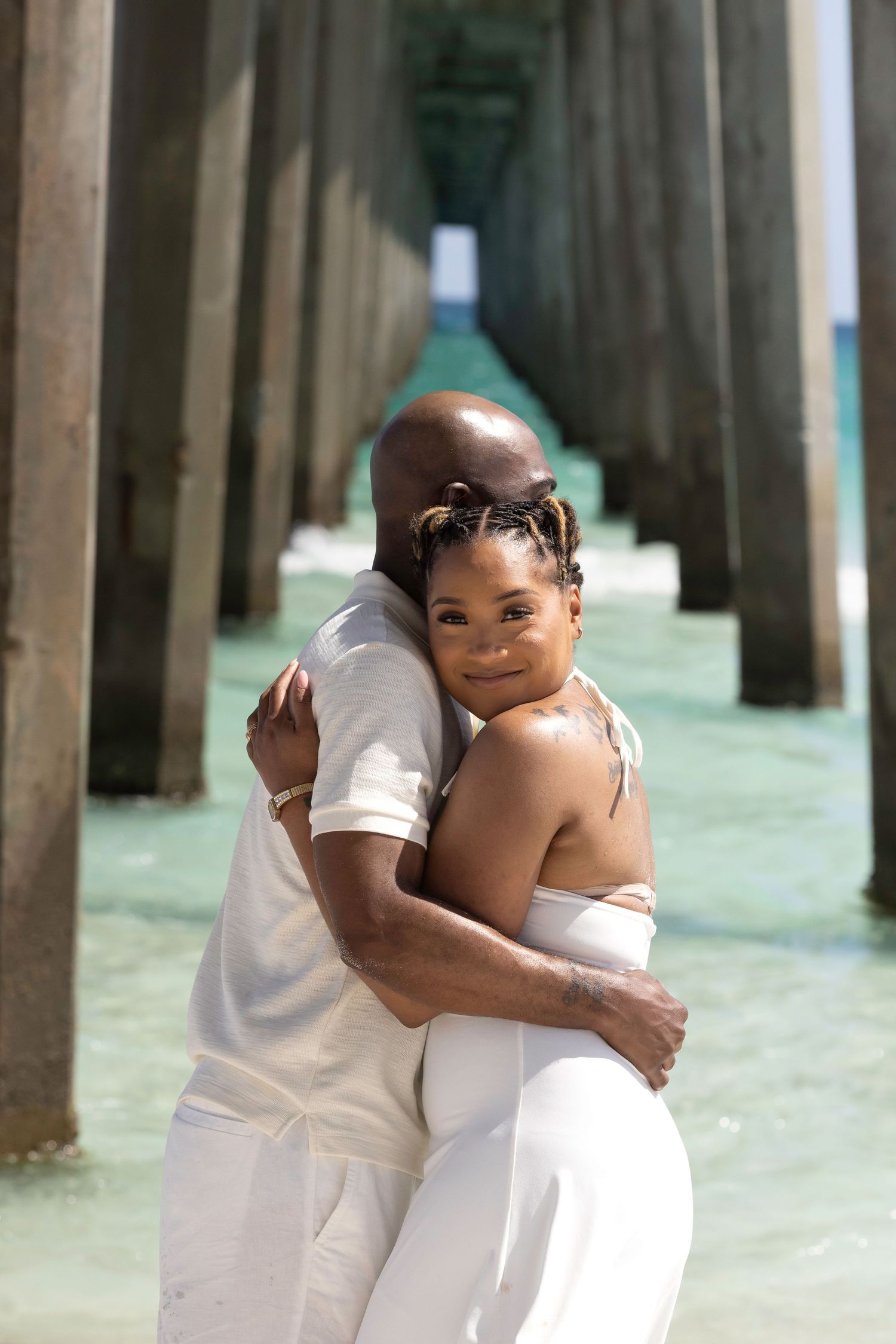 A couple in white embraces under a pier with turquoise water, sharing a romantic beach moment.