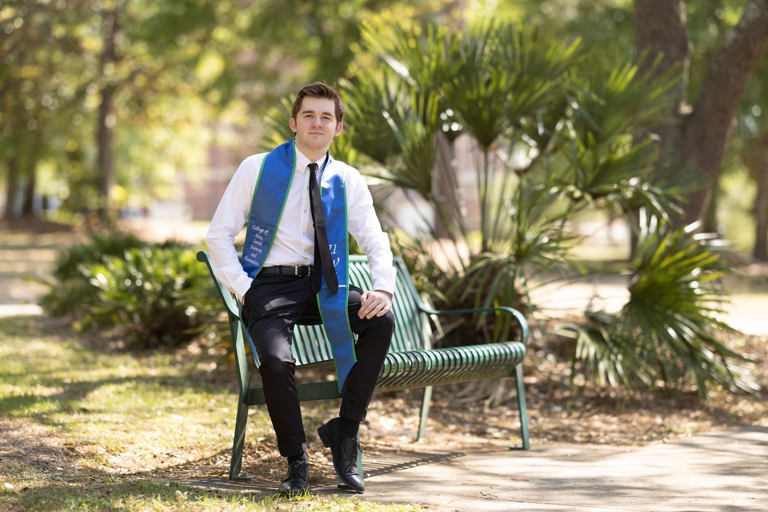 Young man in formal attire sitting on a green park bench surrounded by lush greenery.
