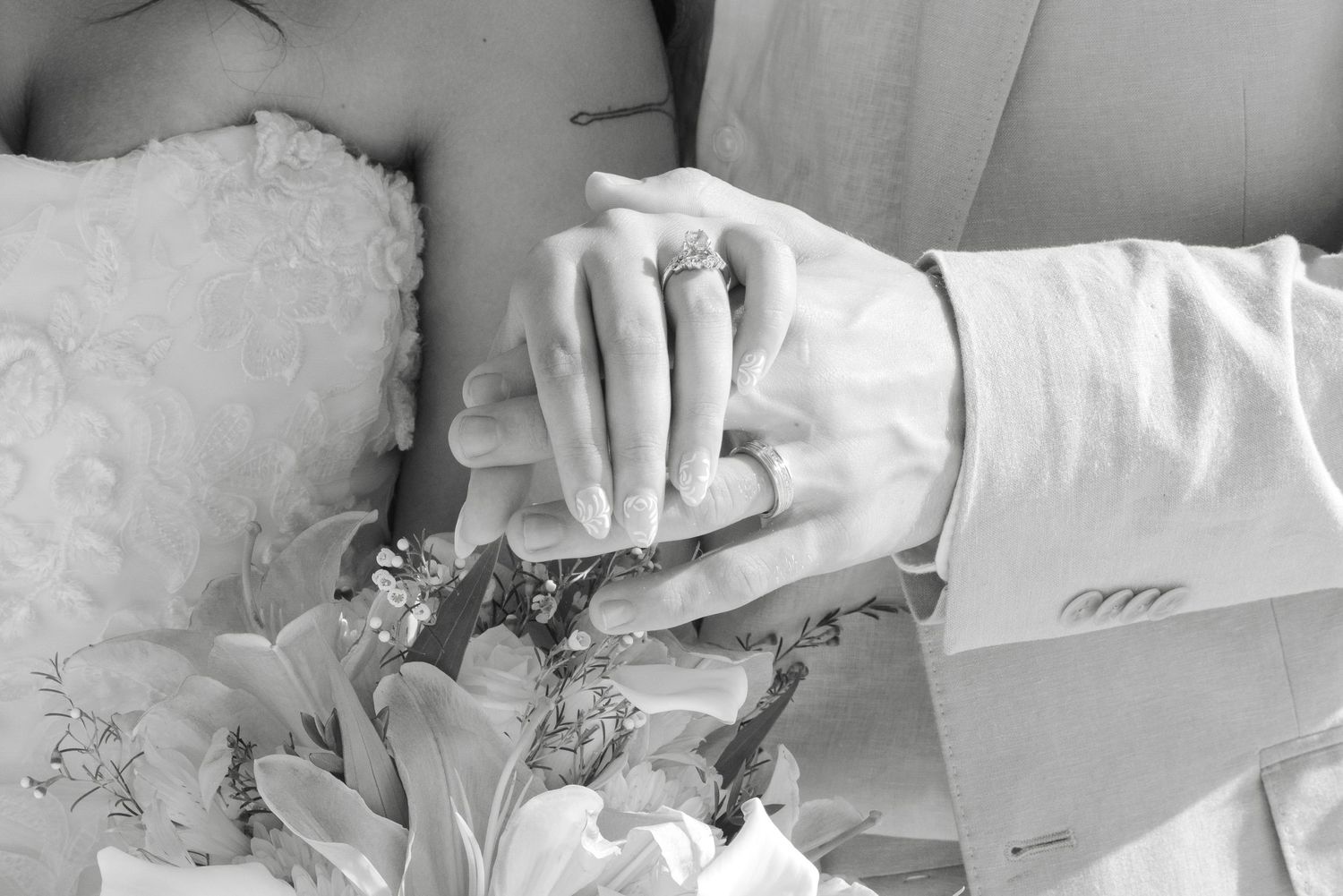 Black and white photo of bride and groom holding hands, showing wedding rings and bouquet of flowers.