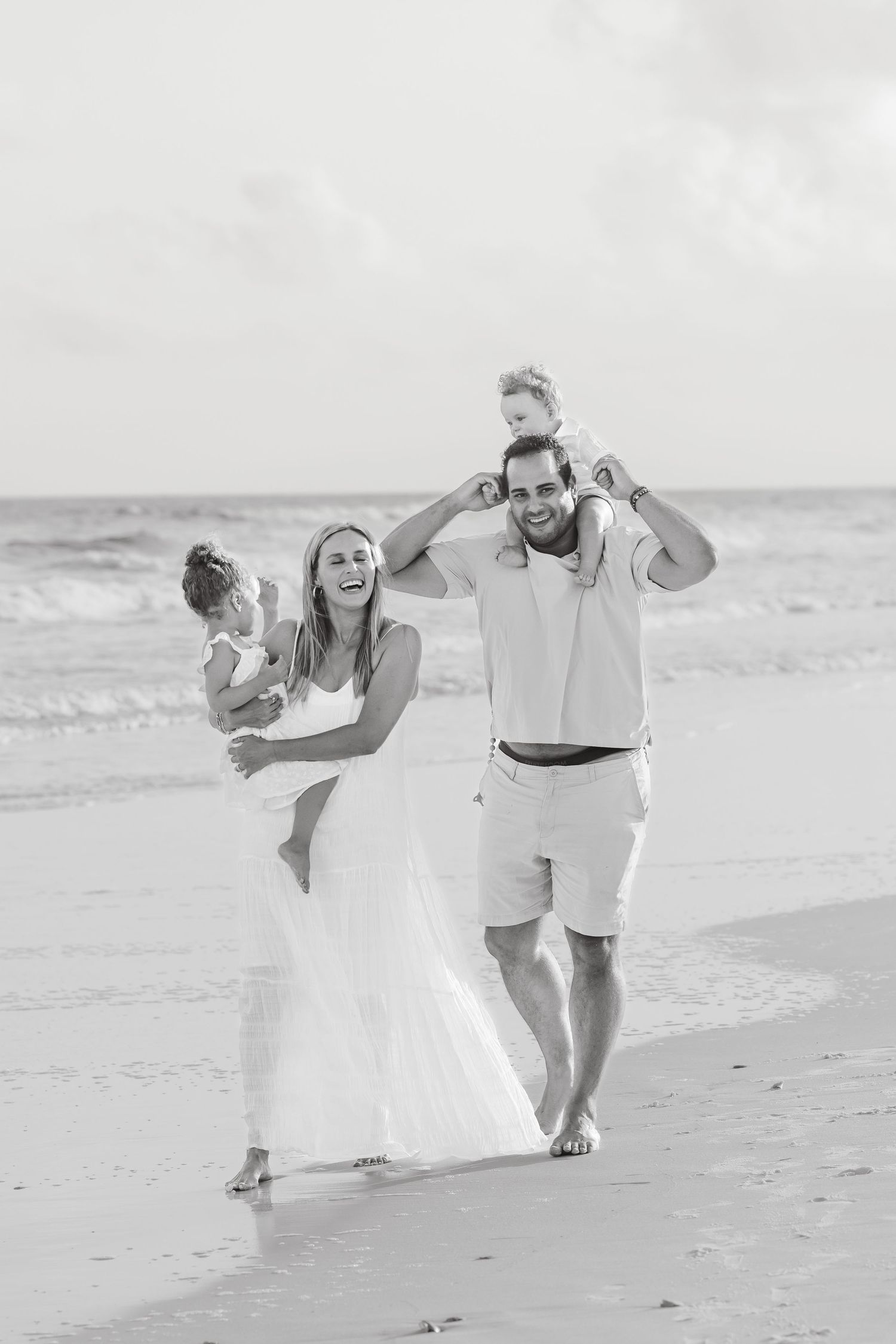 Happy family walking on beach, parents carrying two children, black and white photo, ocean waves in background.