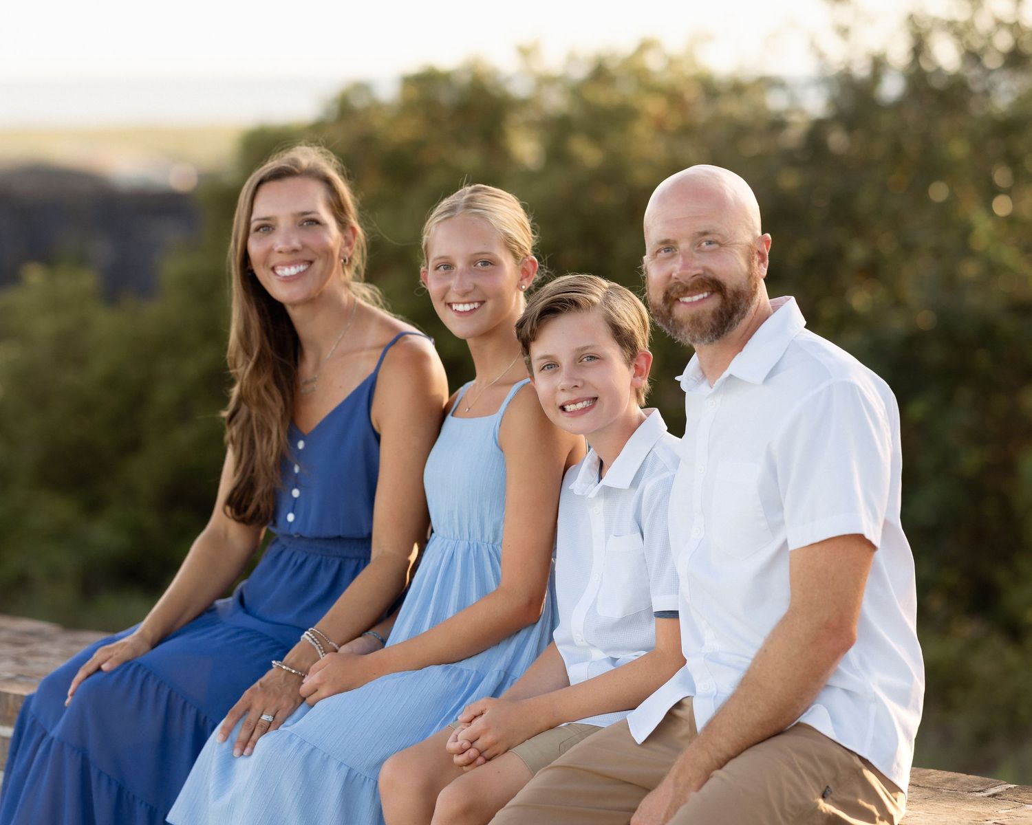 A smiling family of four poses outdoors in blue and white outfits during golden hour.