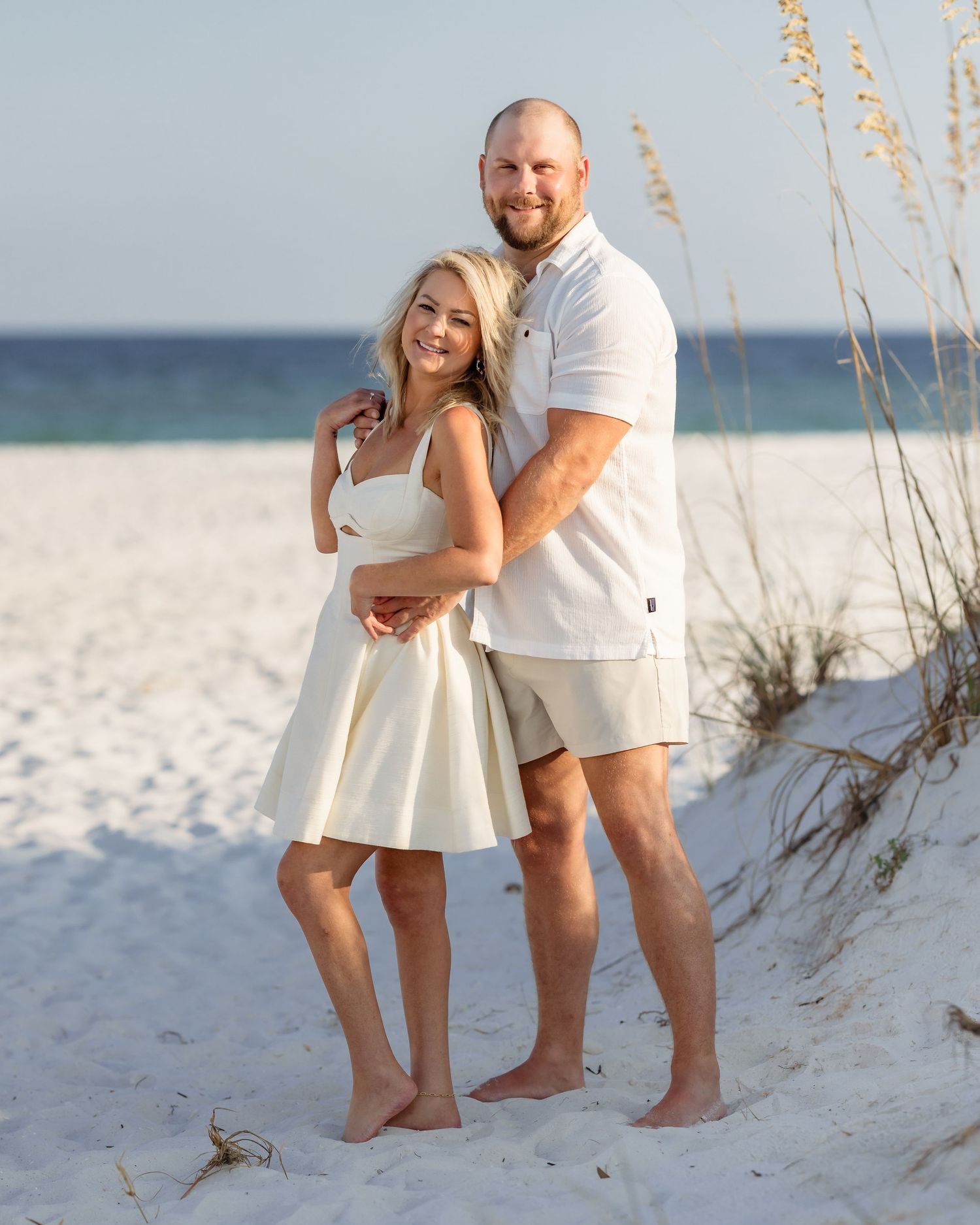 Couple in white outfits posing on sandy beach with sea oats, smiling together near ocean.
