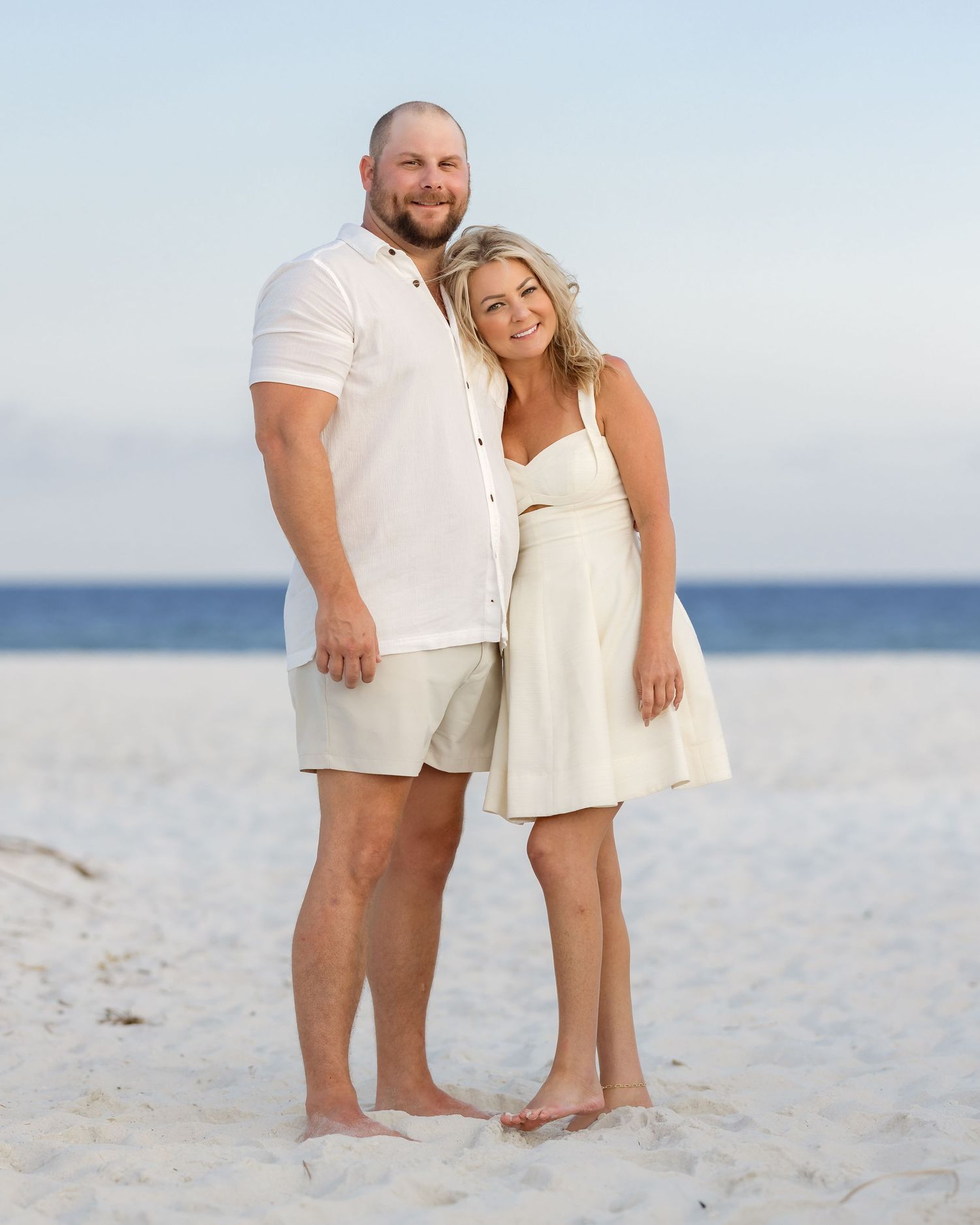 Couple in matching white outfits posing together on a sandy beach with calm ocean waters in the background.