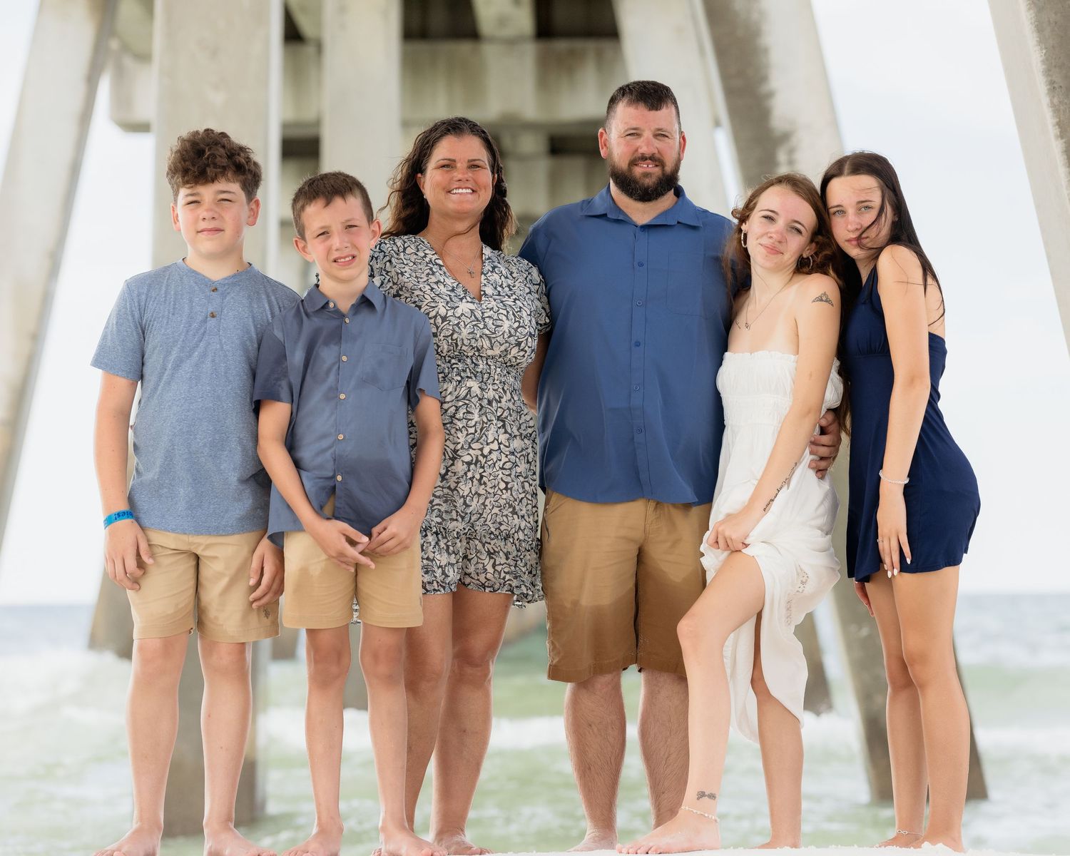 Family of six smiling together on a sunny beach near a pier, wearing casual summer outfits.
