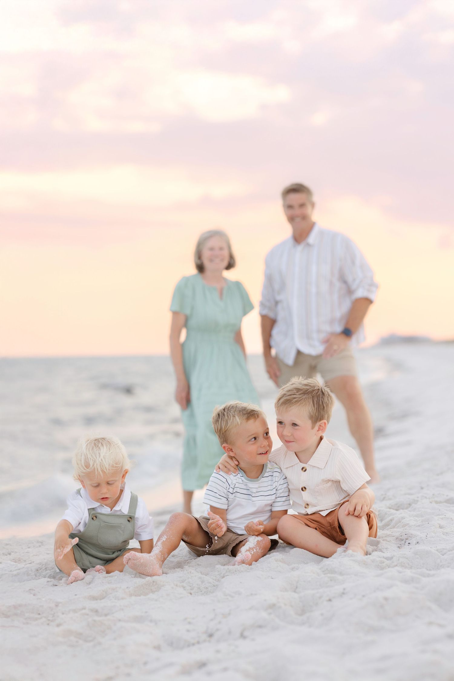 Children laying playfully in soft white sand with family in background during sunset.
