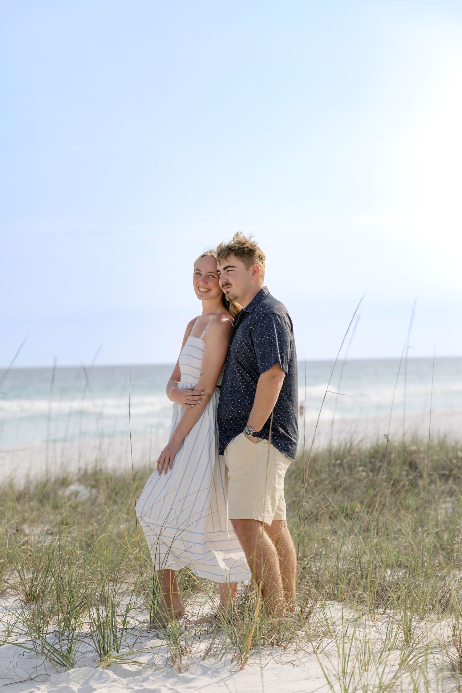 A couple embraces on a sunny beach, woman in white dress, man in navy shirt and khaki shorts, with ocean in background.