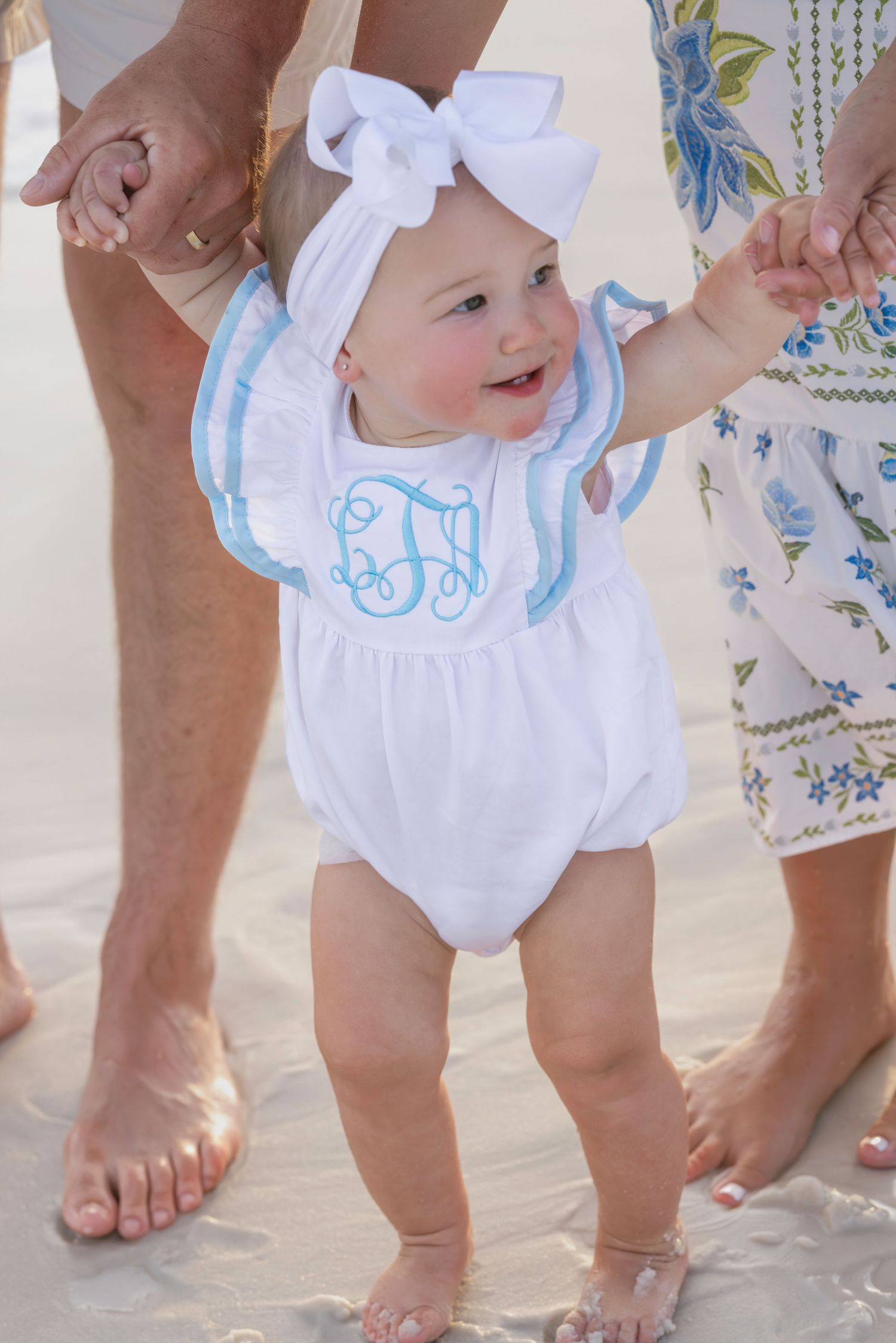 Smiling baby in white monogrammed romper and bow headband stands on sandy beach held by parents.