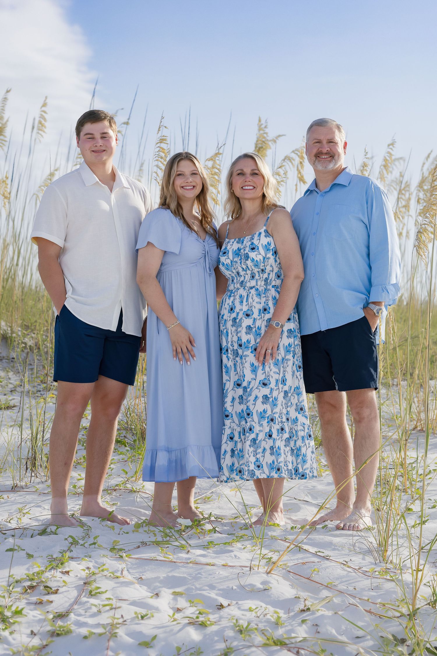 Happy family of four posing on sandy beach dunes, wearing coordinated blue and white outfits.