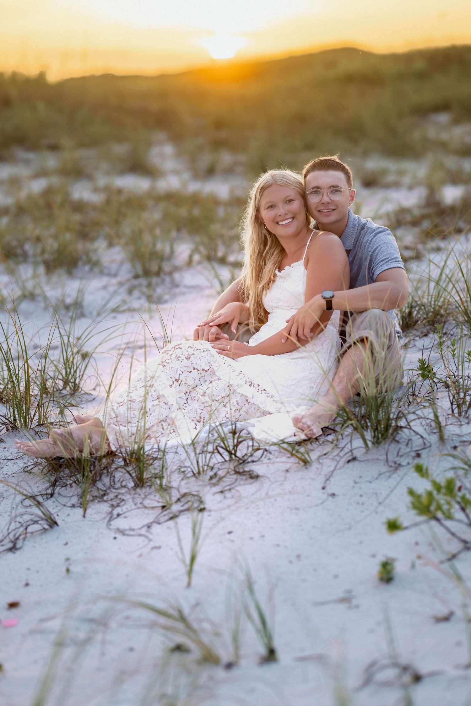Couple sitting together on white sand dunes at sunset, smiling, woman in white dress, man in gray shirt, golden hour.