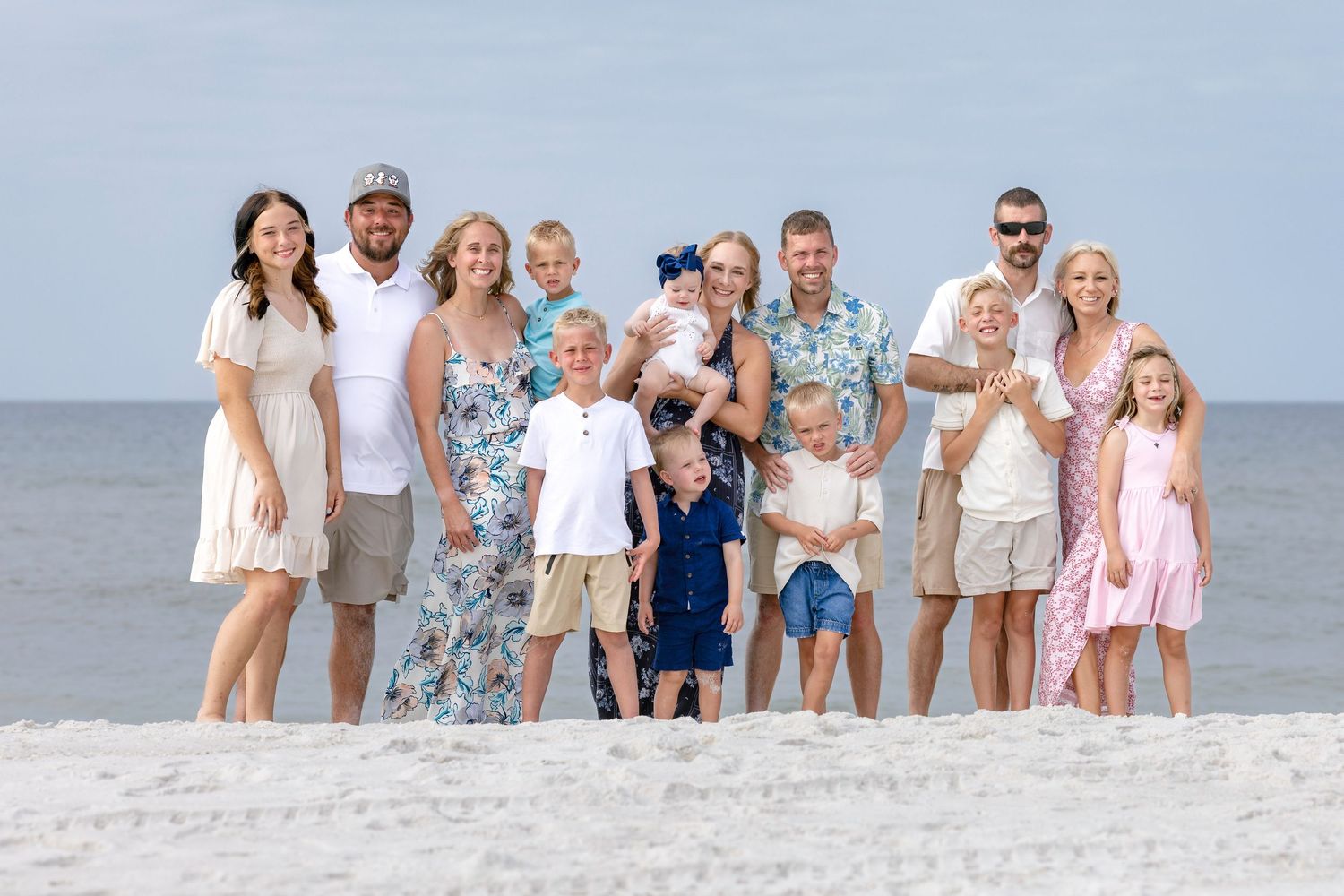 Large family group smiling together on a sunny beach, wearing coordinated outfits in white, blue, and pink tones.