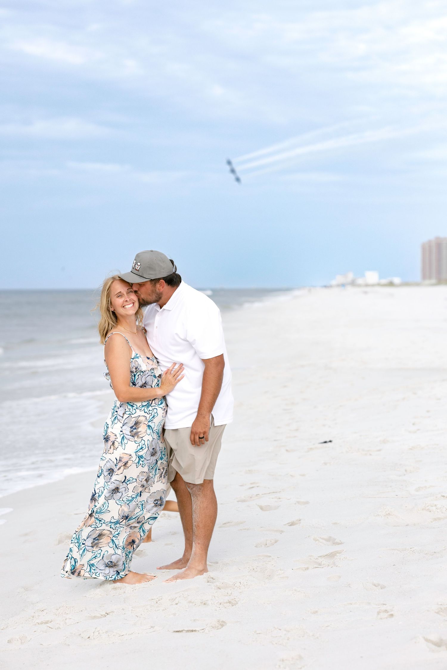 Couple embracing on white sand beach with airshow planes in background, woman in floral dress, man in white shirt.