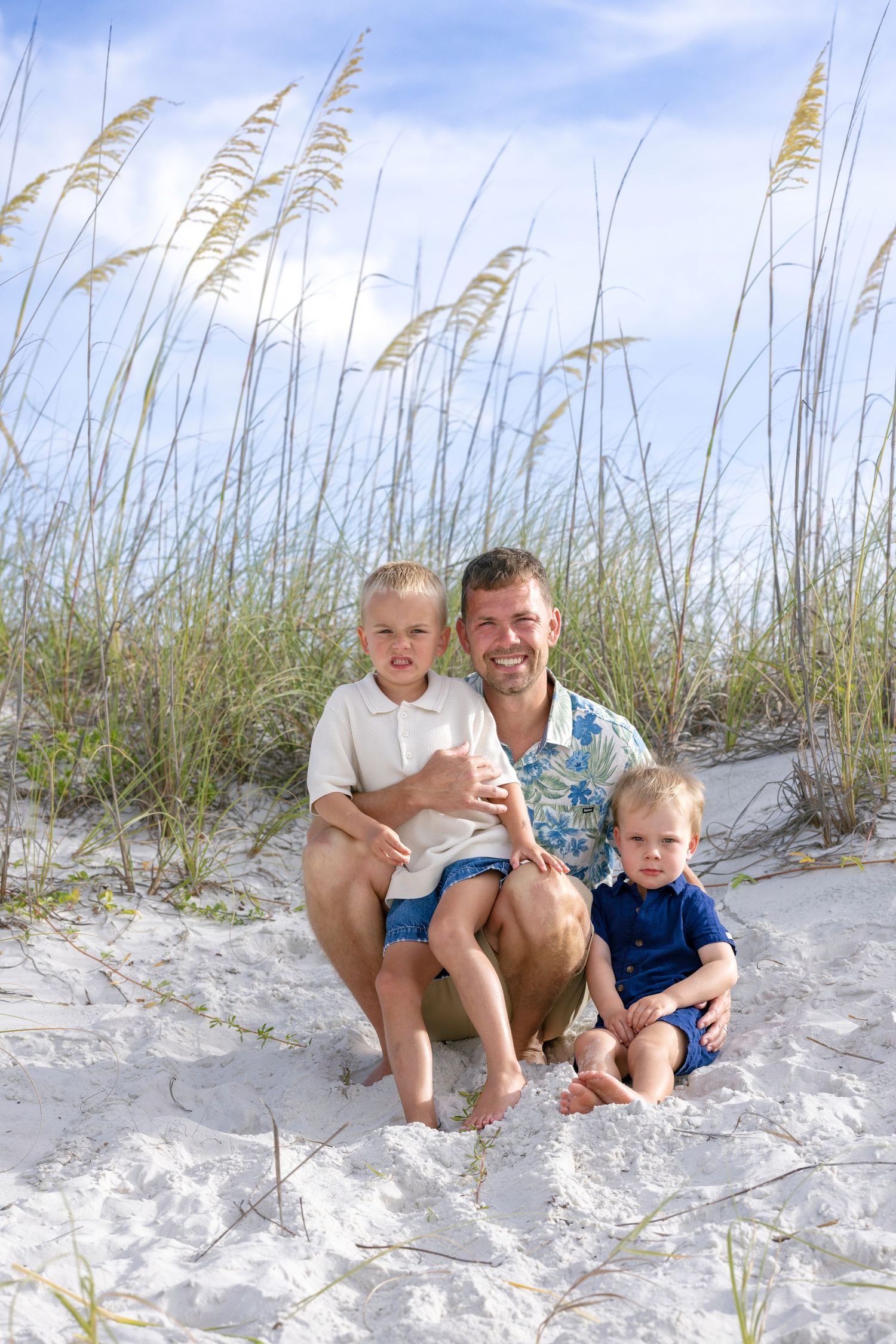 Father sitting in beach dunes with two young sons, smiling among tall sea oats.