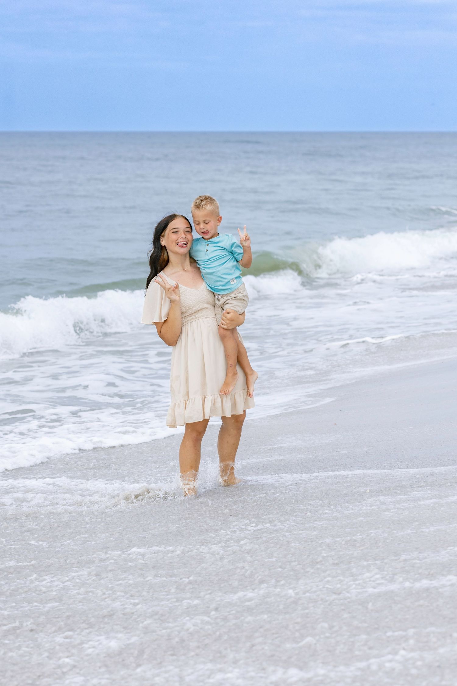 Smiling mother in cream dress holding toddler boy on sunny beach with ocean waves behind them.