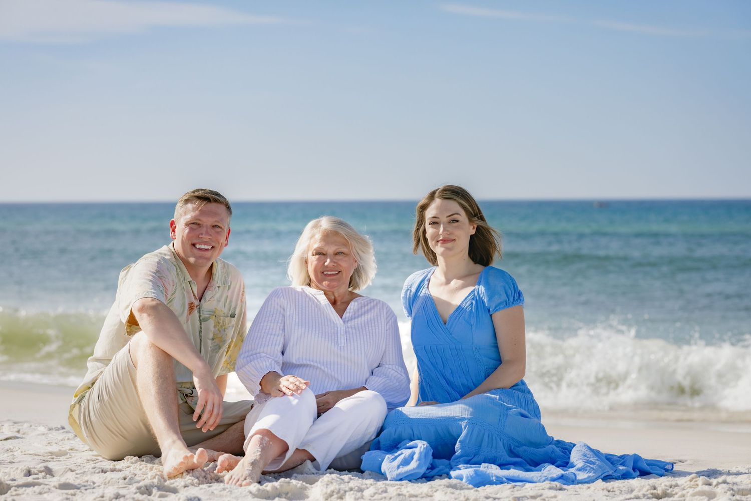 Three people smiling and sitting together on a sandy beach with ocean waves in the background on a sunny day.