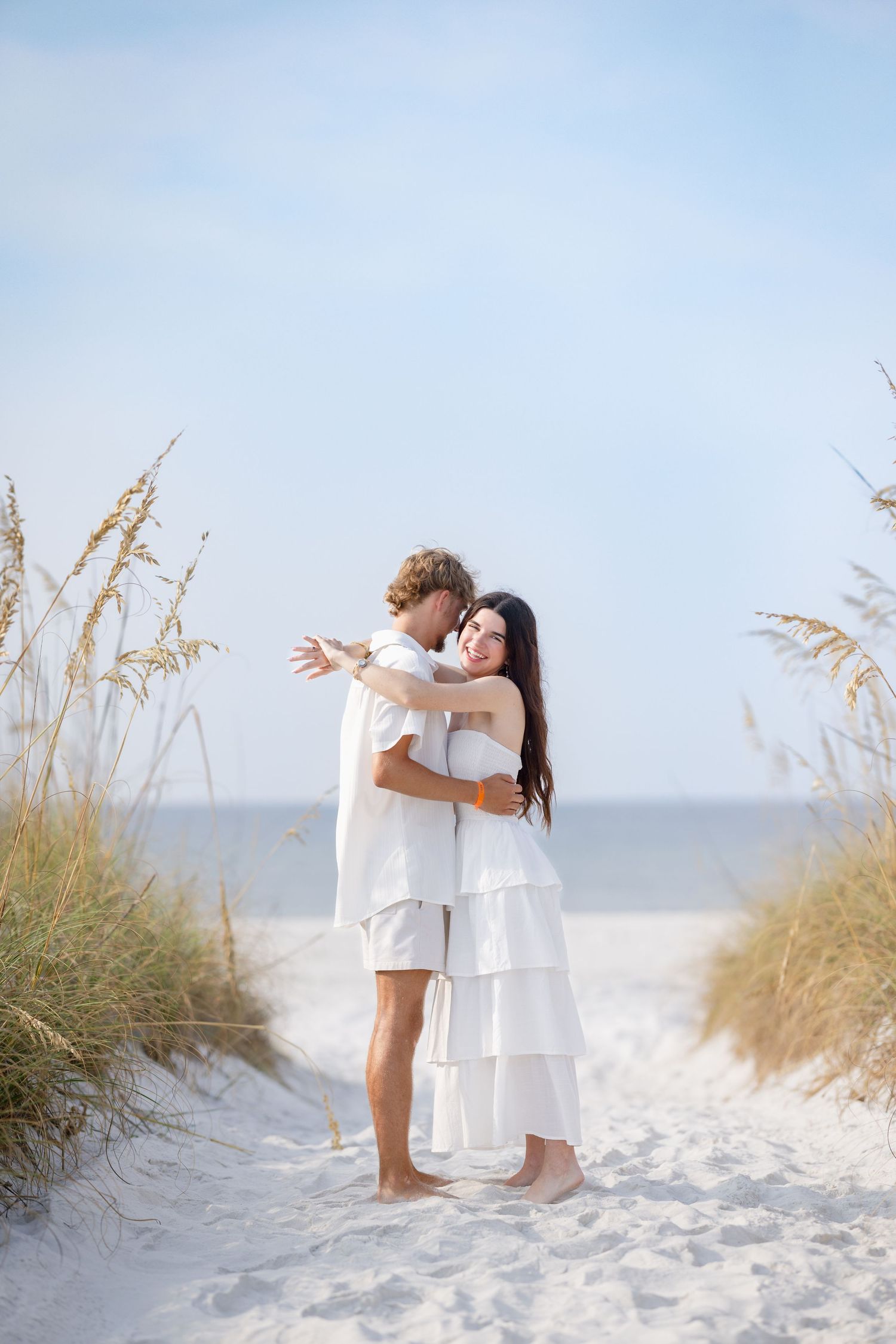 Couple embracing in white outfits on sandy beach path surrounded by sea oats.