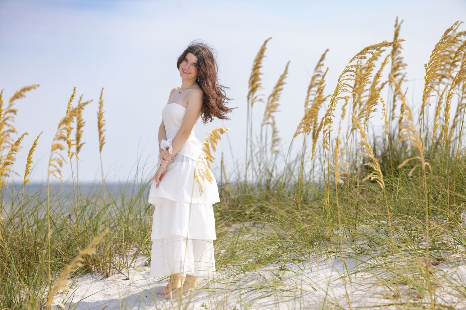 Woman in white tiered dress standing on beach dunes surrounded by tall sea oats grass.