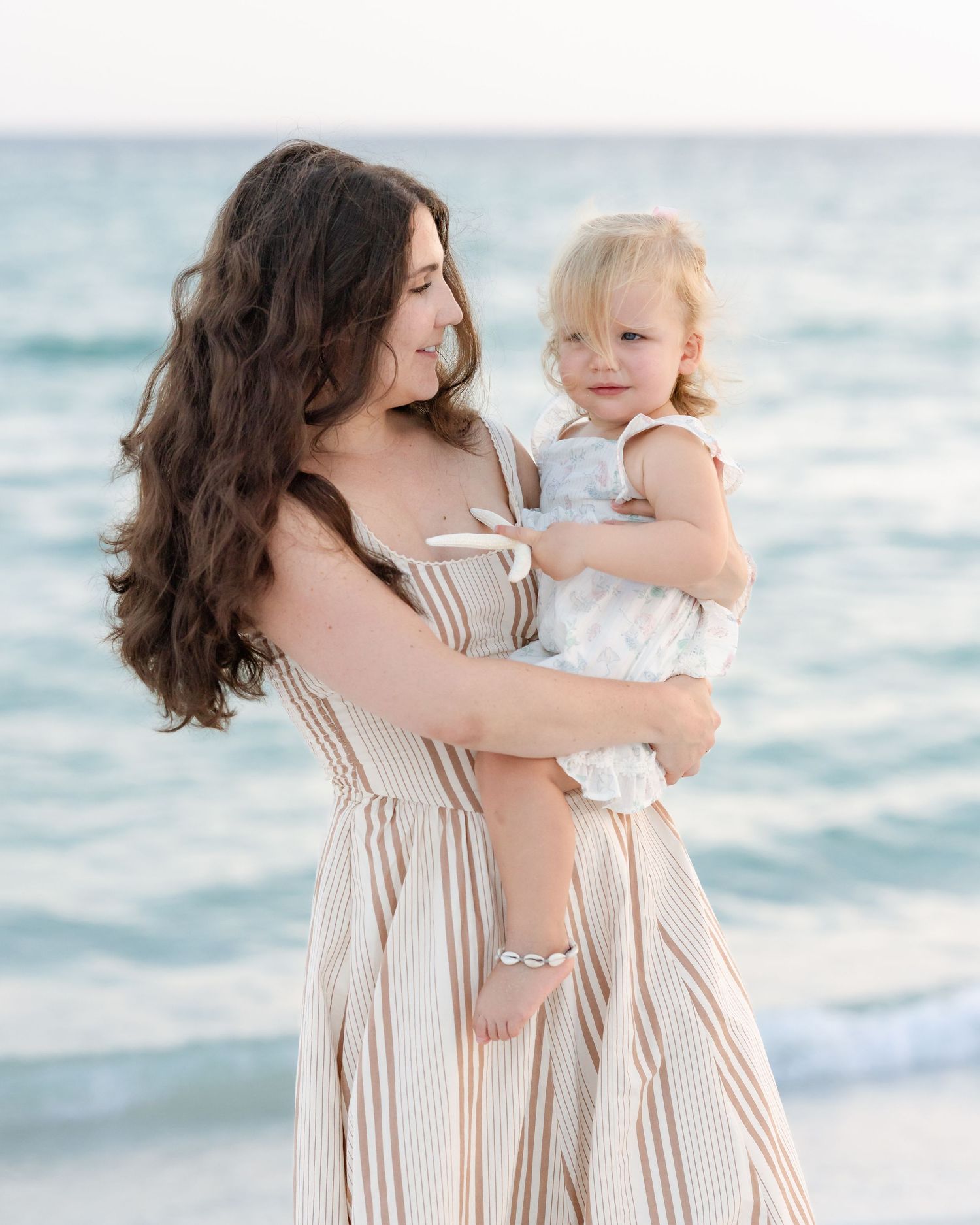 Brunette mother in striped dress holds blonde toddler girl at beach with turquoise ocean in background.