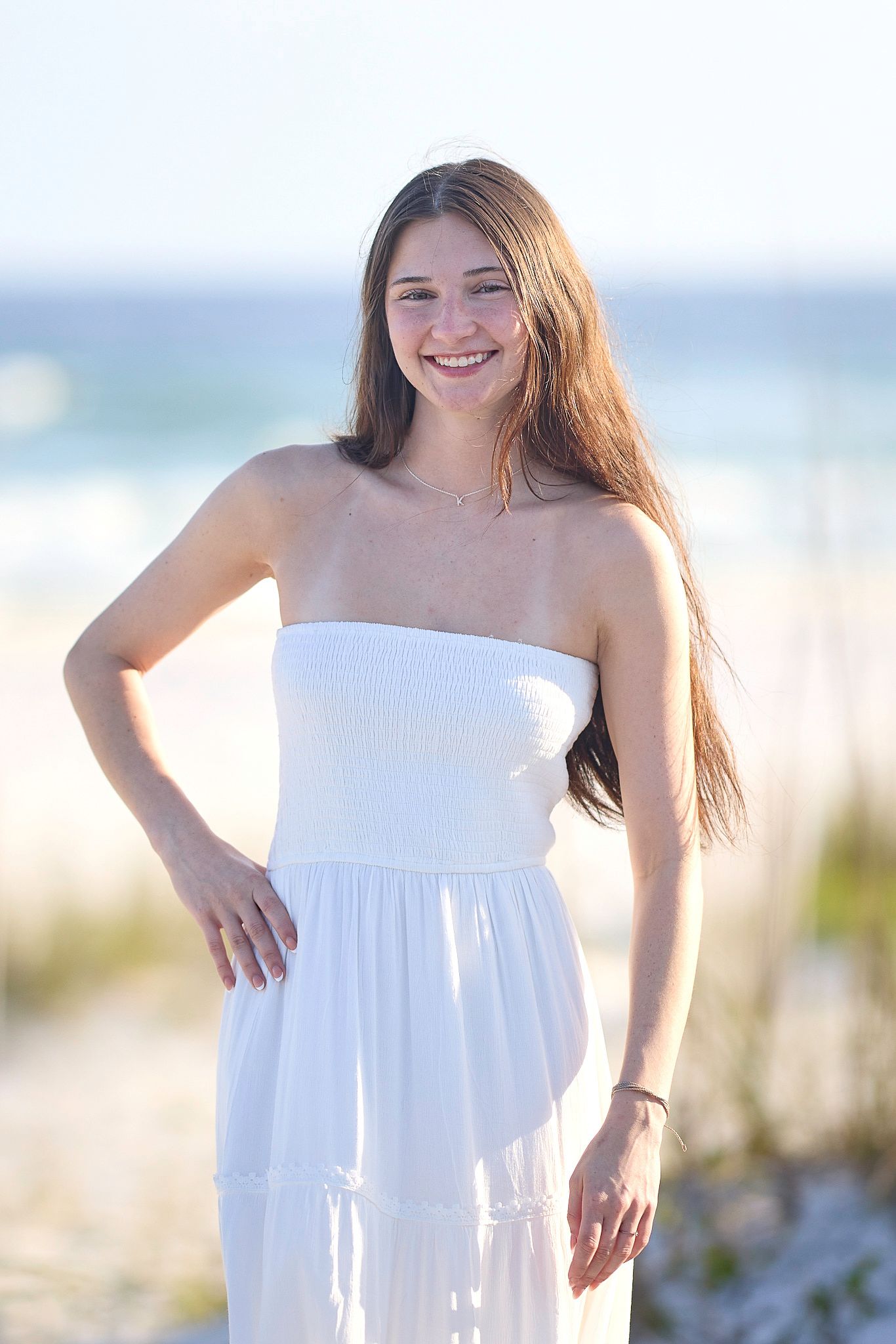 Someone in a strapless white dress stands playfully on a sunny beach with soft lighting and ocean views.