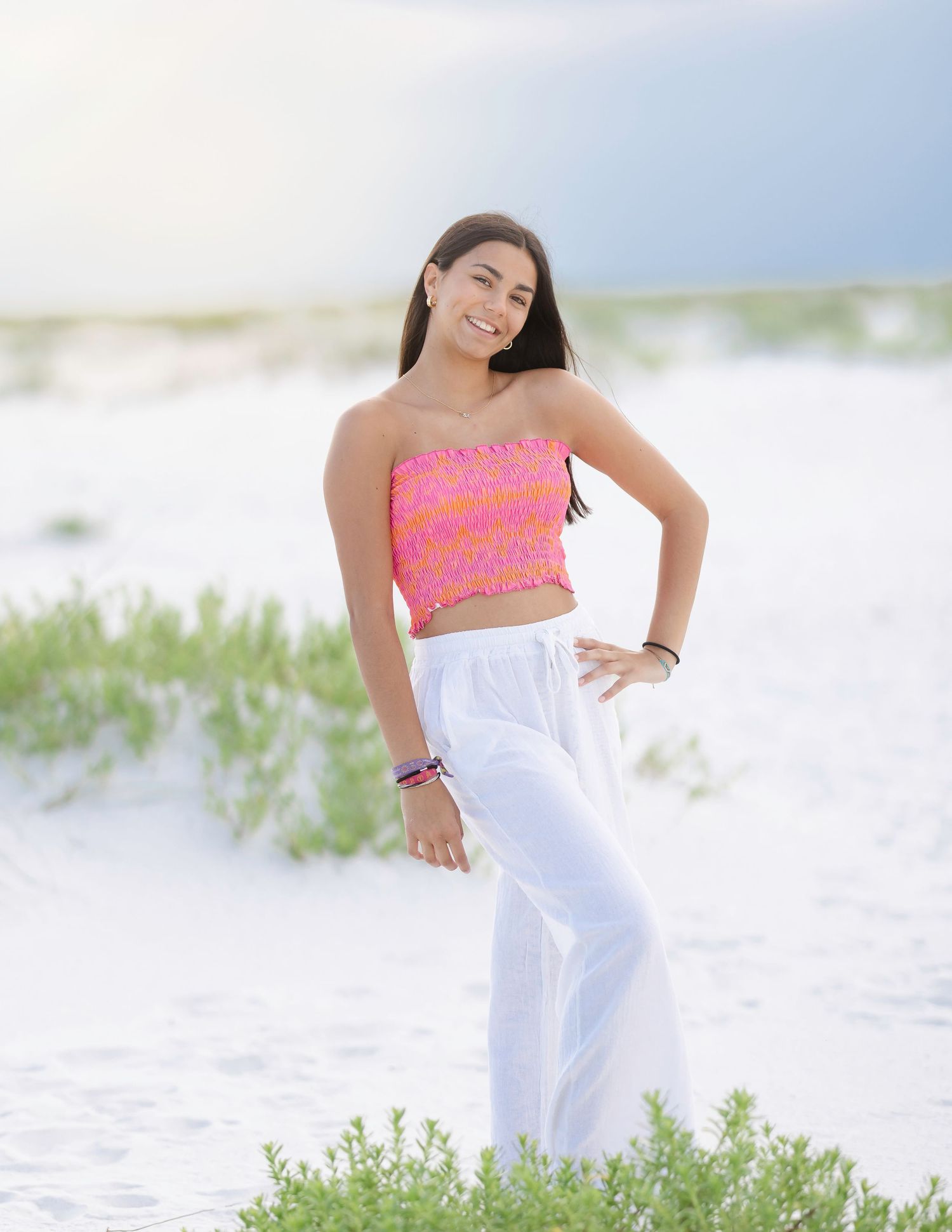 Woman in coral top and white pants dances on sandy beach near ocean waves.