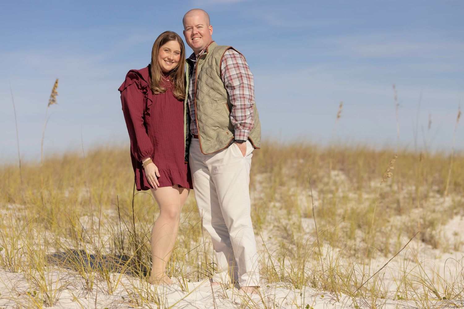A couple walks together on a sandy beach path through tall grass and dunes on a sunny day.