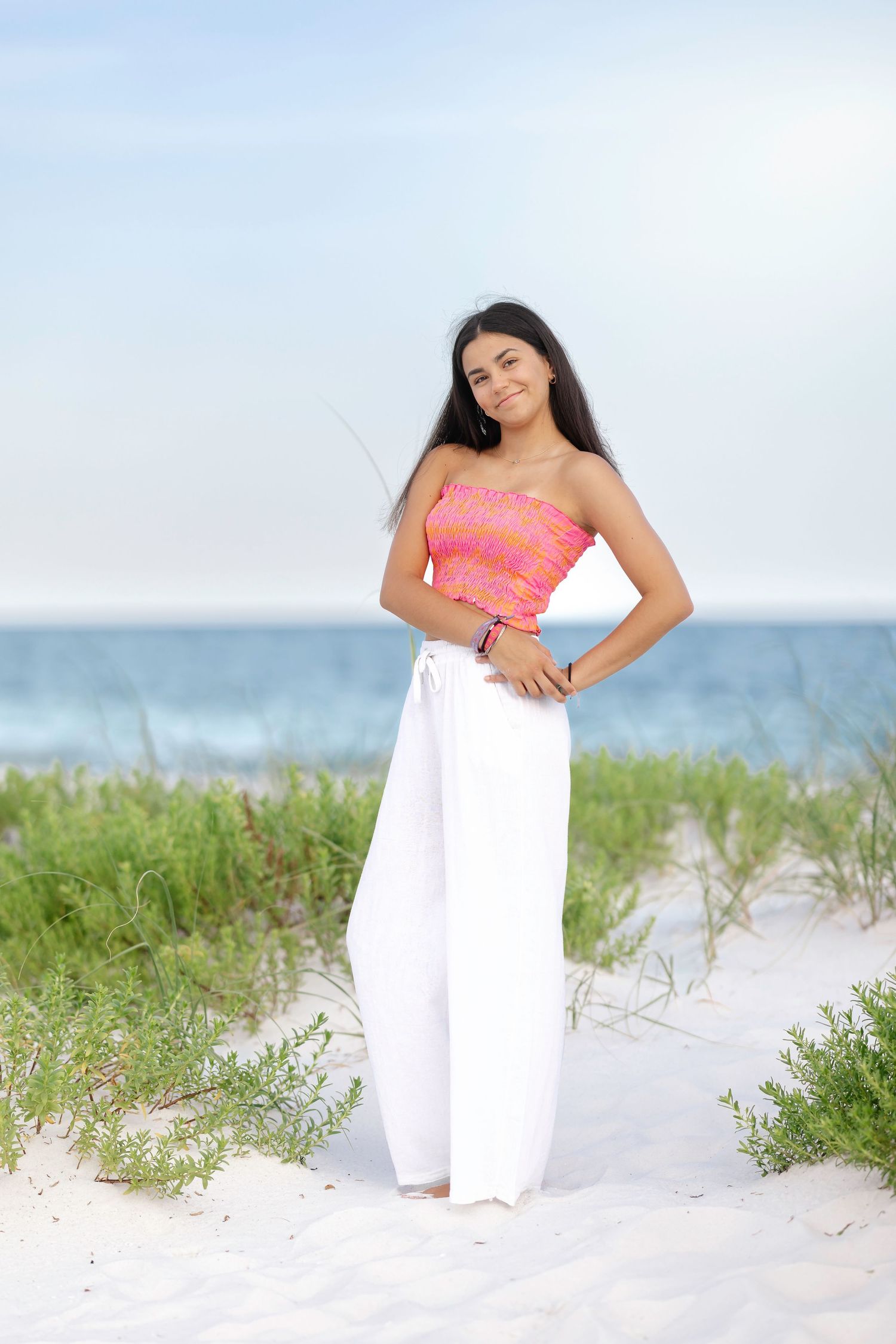 A beachside photo session featuring white flowy pants and coral top against sand dunes and ocean backdrop.