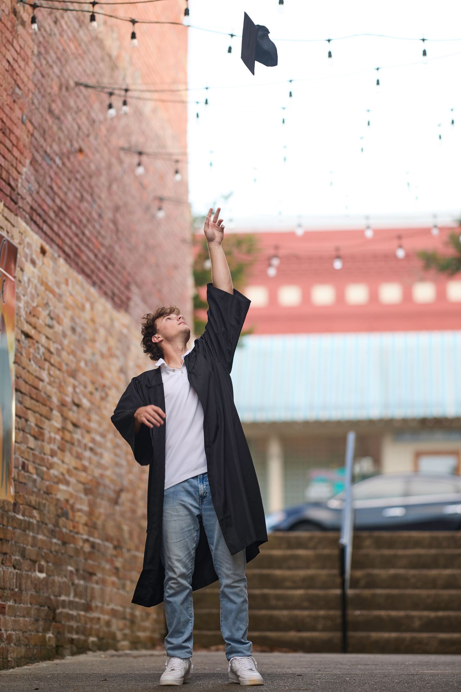 A graduate celebrates by tossing their cap in the air against a brick wall backdrop with string lights overhead.