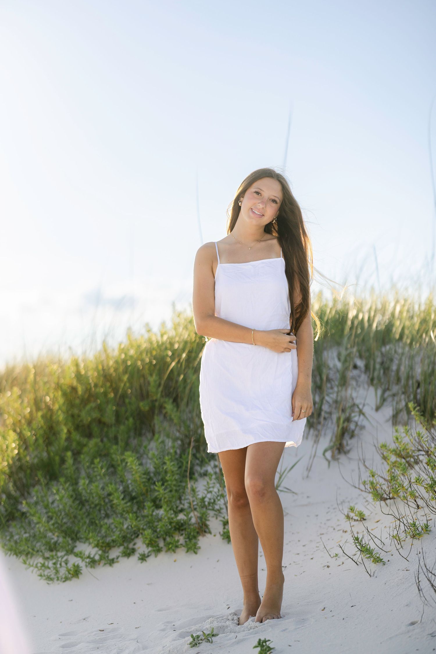 Woman in white dress stands playfully on sandy beach with coastal vegetation.