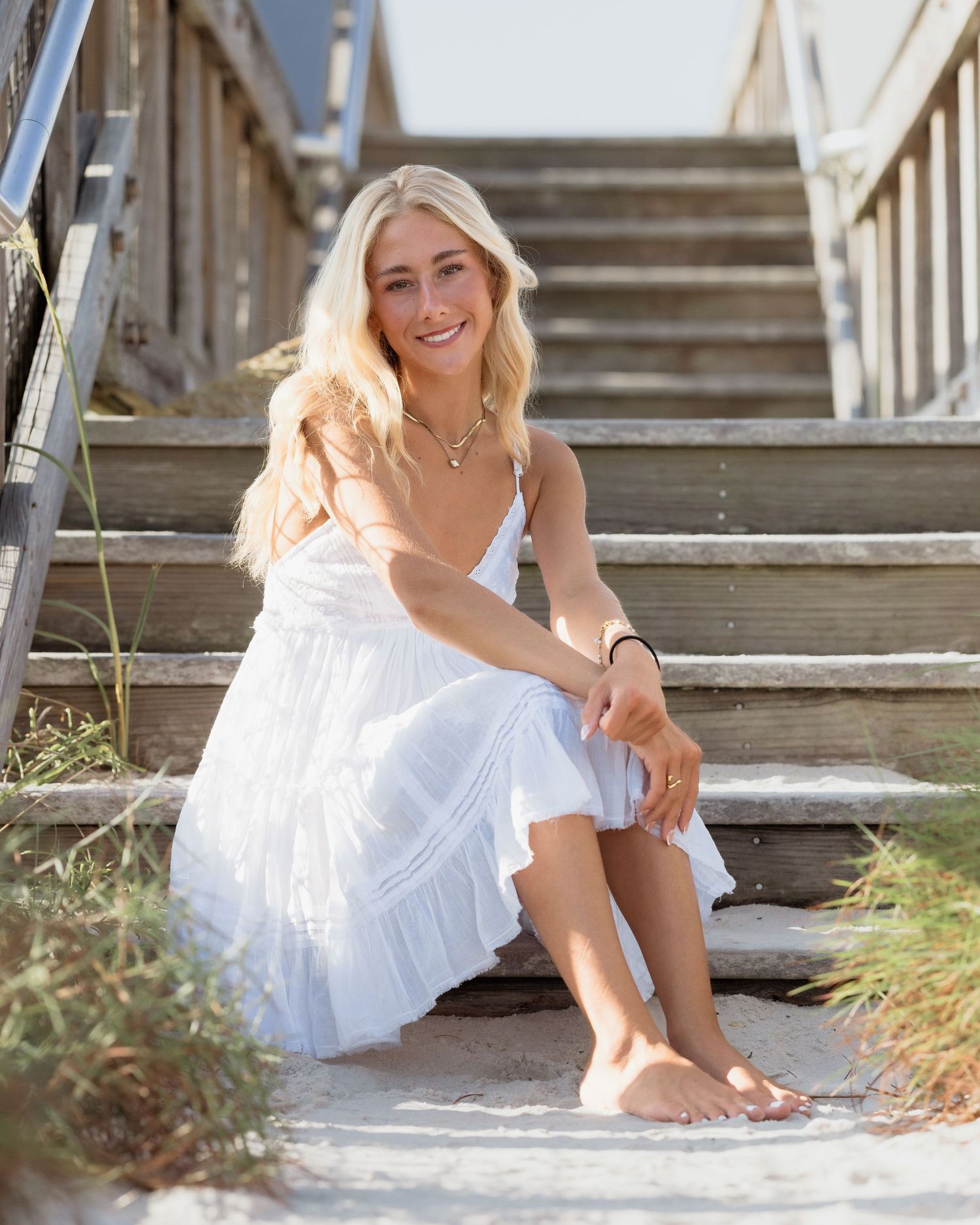 Smiling blonde woman in white sundress sitting barefoot on wooden beach stairs in sunny outdoor setting.