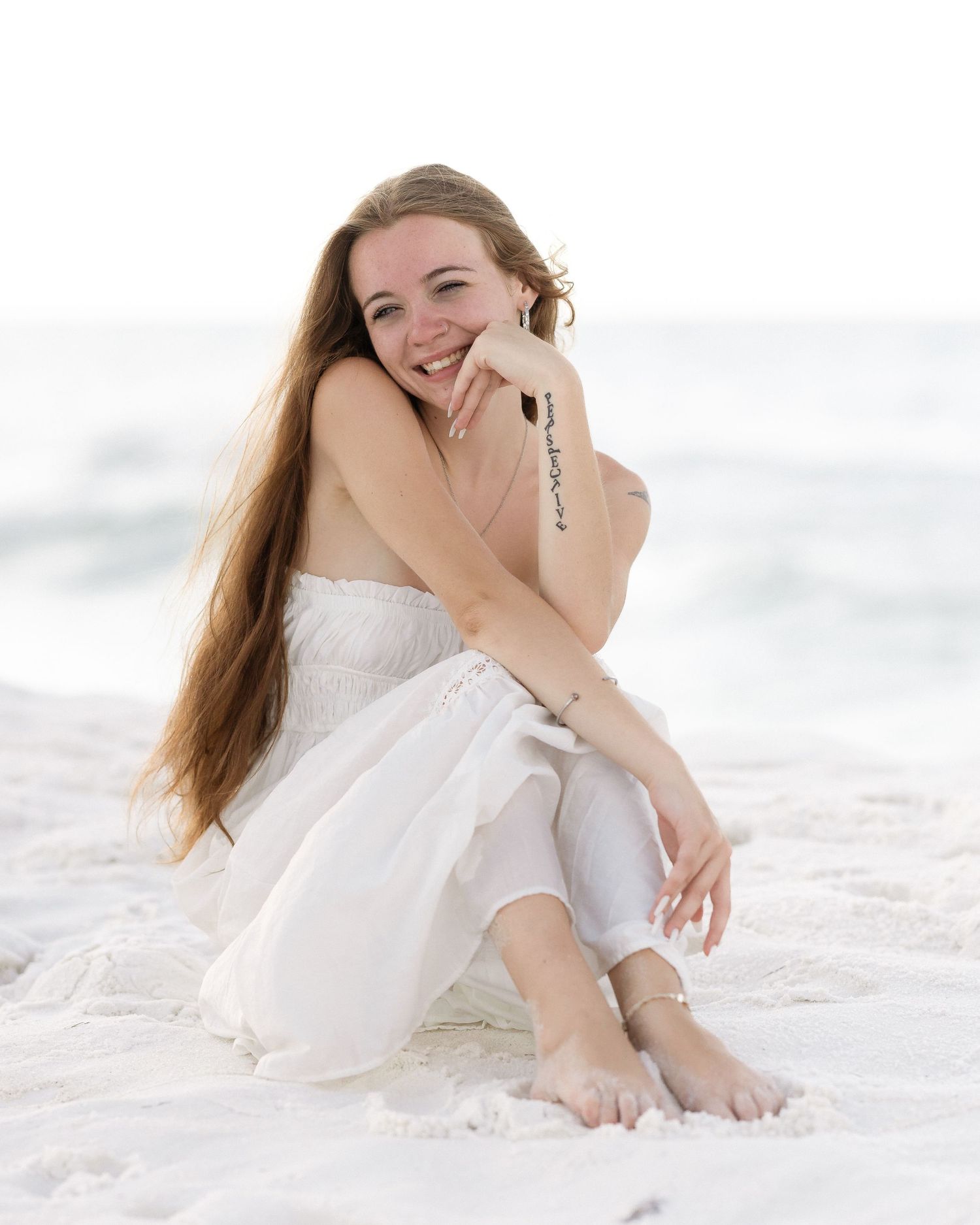 Young woman in white dress sitting barefoot on sandy beach, smiling with long auburn hair.