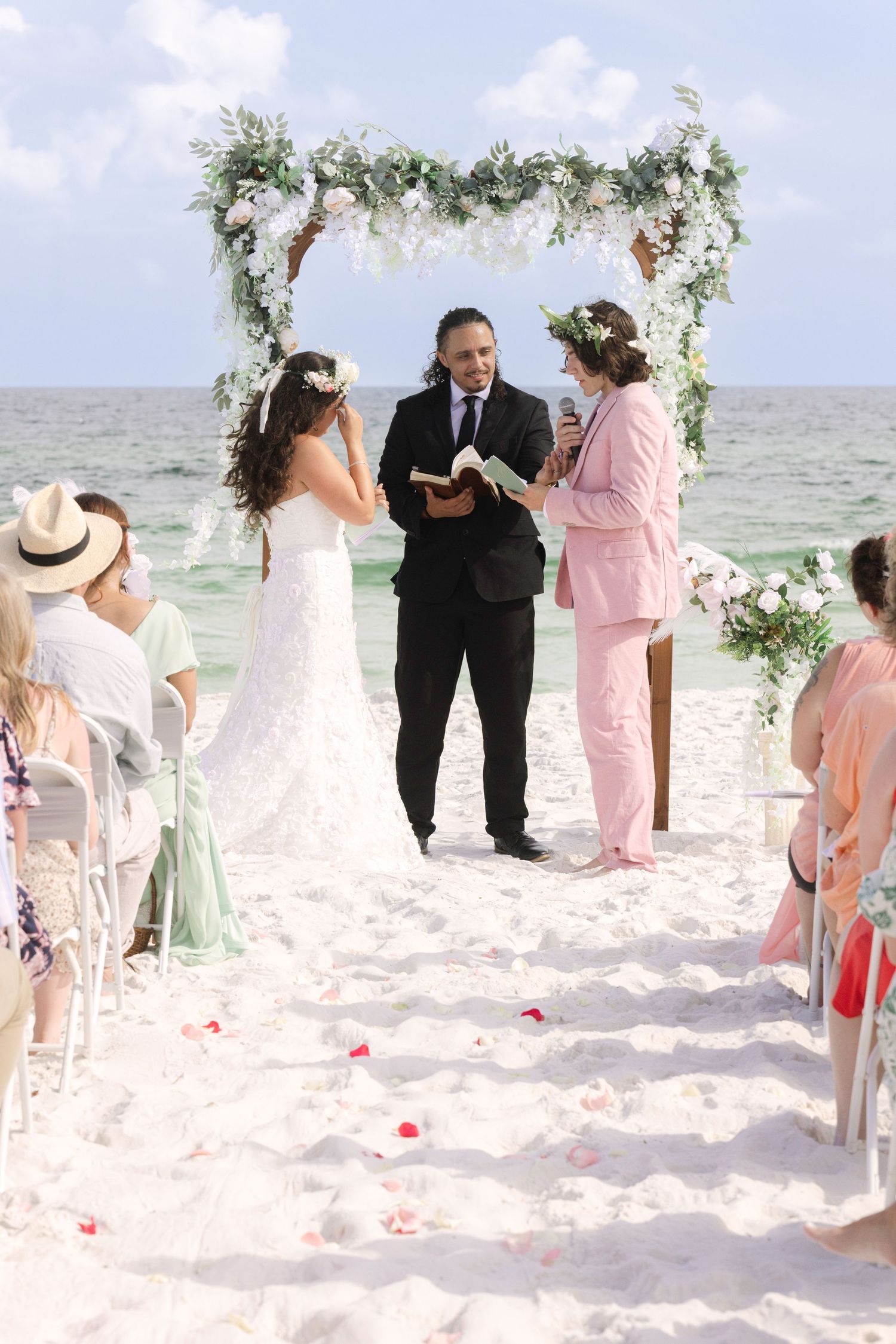 Beach wedding ceremony with bride, groom in pink suit, and officiant under floral arch by the ocean.