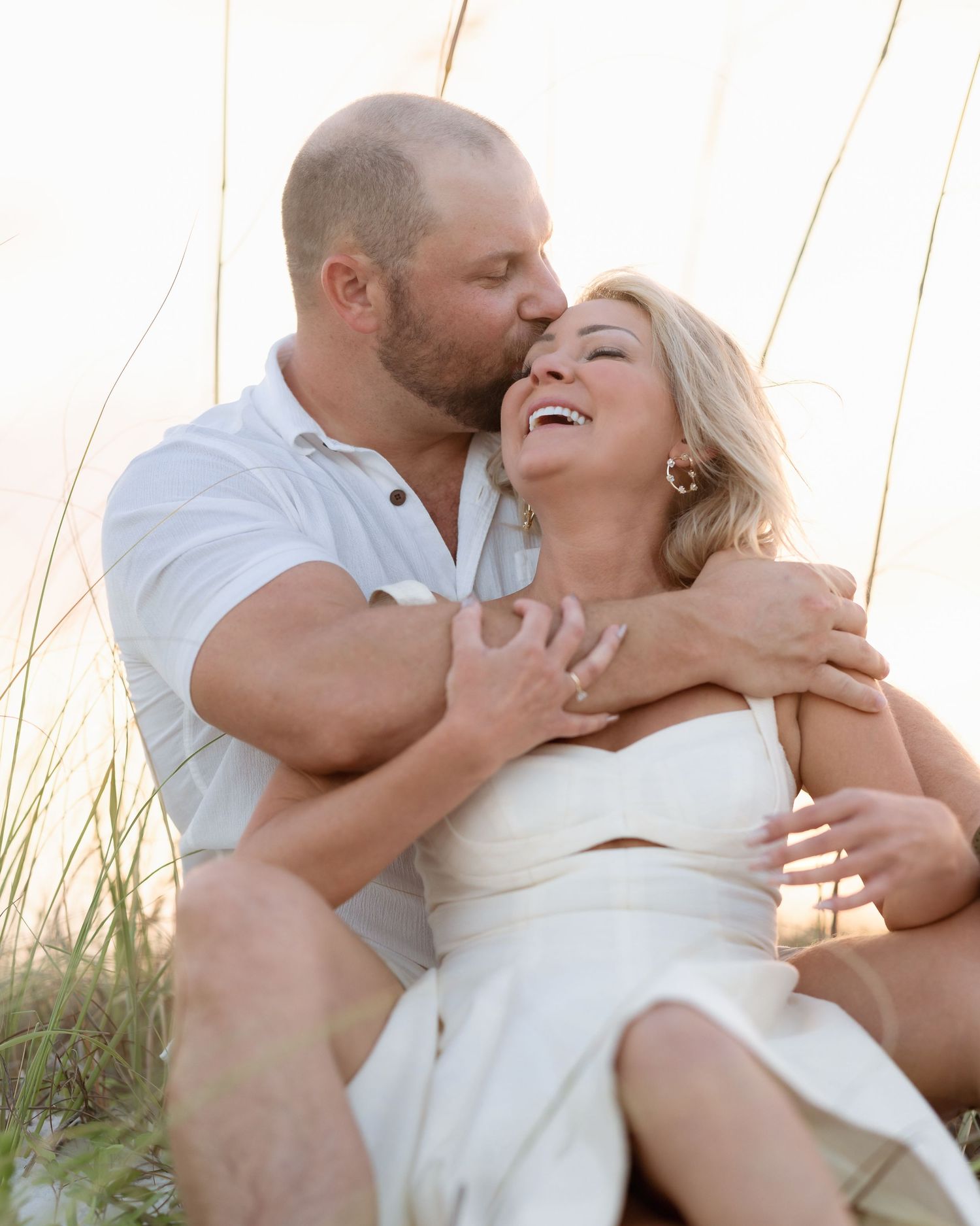 A couple shares a romantic kiss and laugh together in beach dune grass.