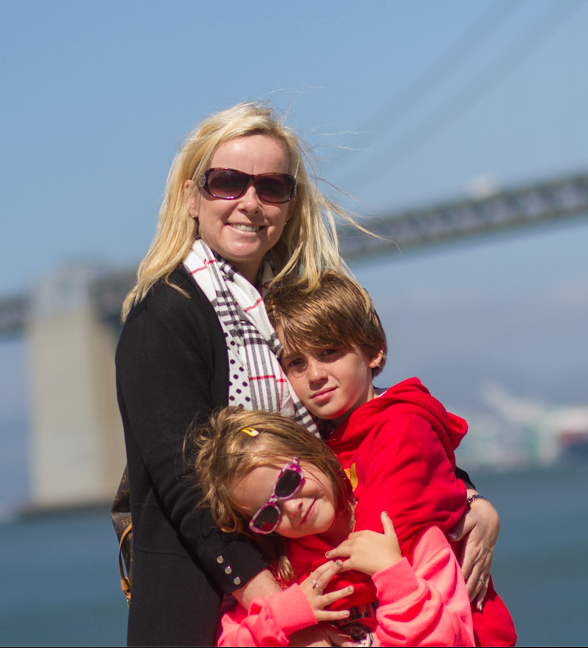 A smiling blonde woman with sunglasses hugs two young children near a waterfront bridge on a sunny day.