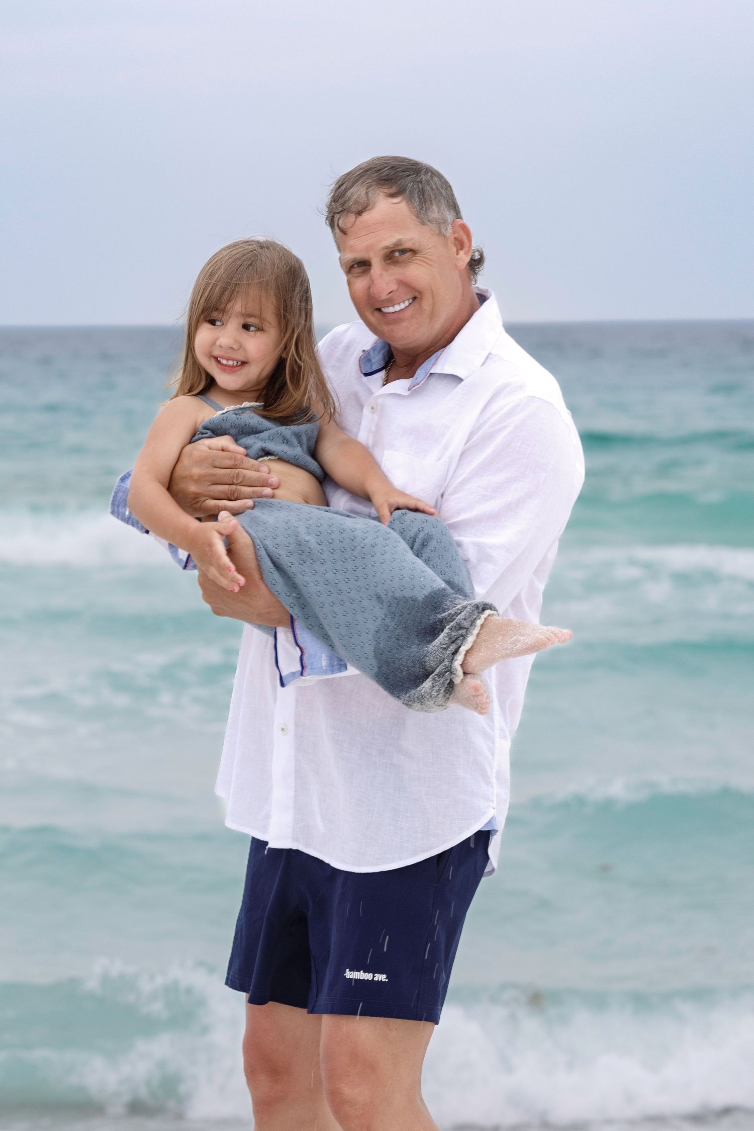 A smiling man holds a young girl wrapped in a towel on a beach with ocean waves in the background.