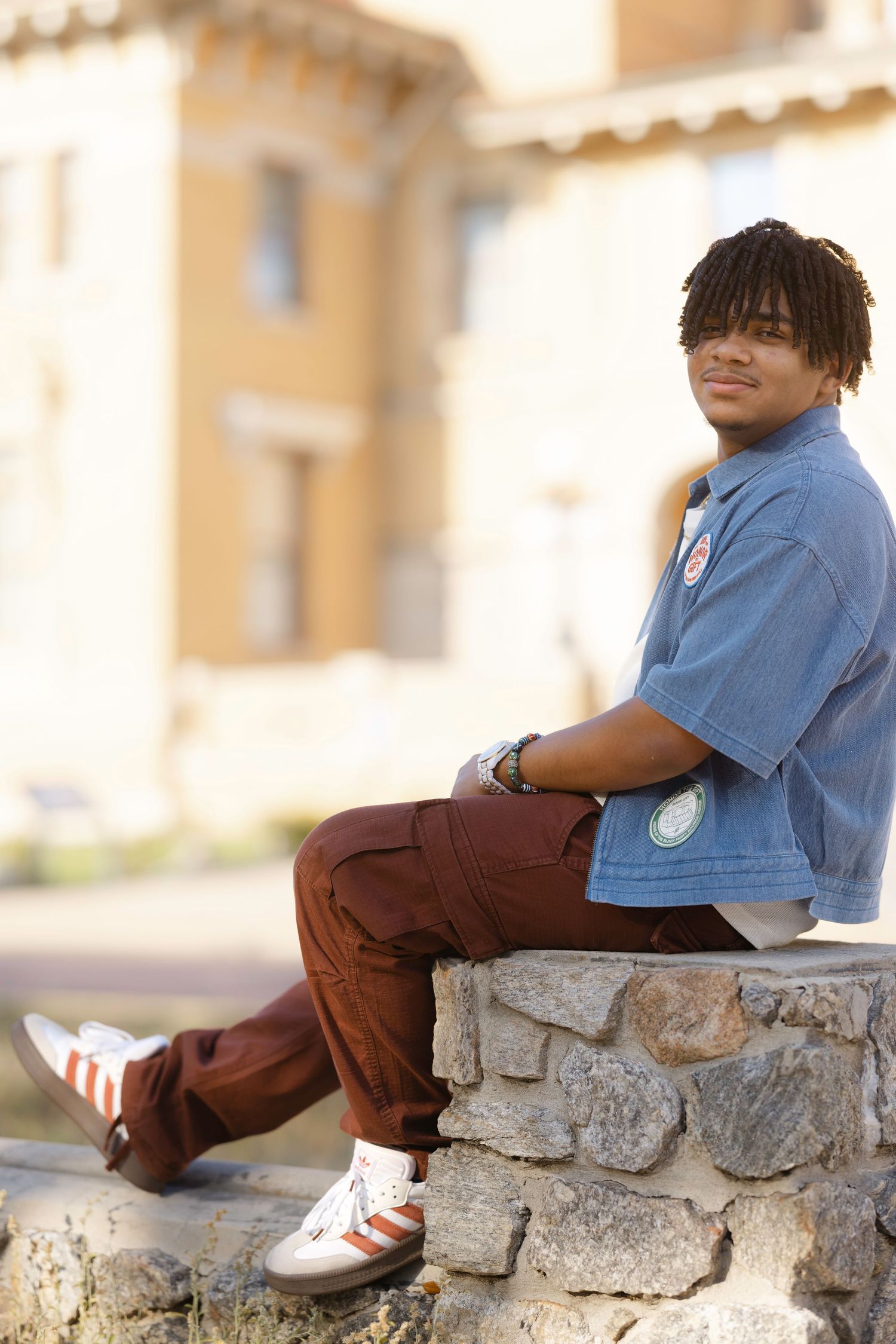 Young man with dreadlocks sits on stone wall wearing blue shirt, brown pants, and Adidas sneakers outdoors.