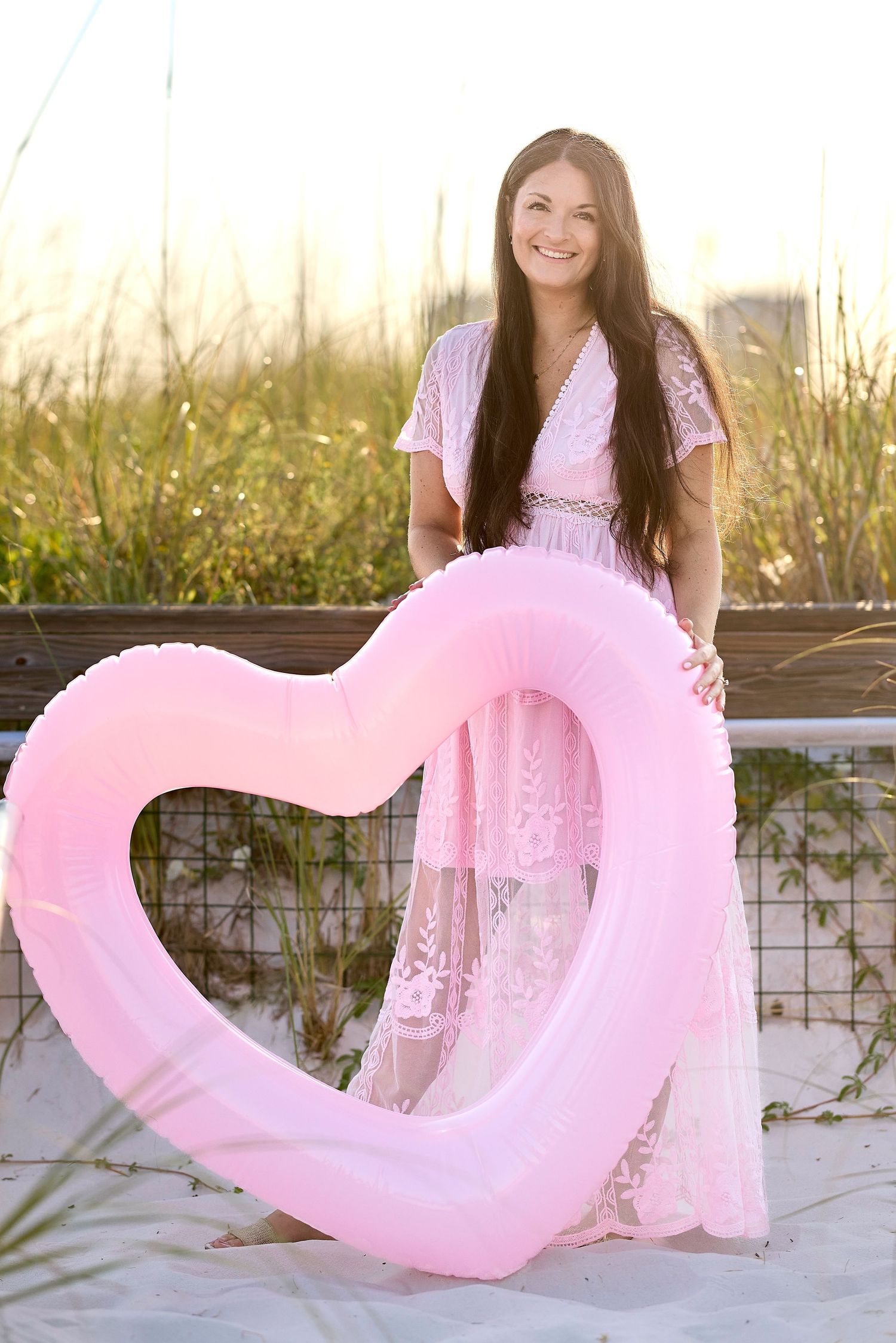 Happy brunette woman in pink lace maxi dress holding heart-shaped inflatable ring on a sandy beach.