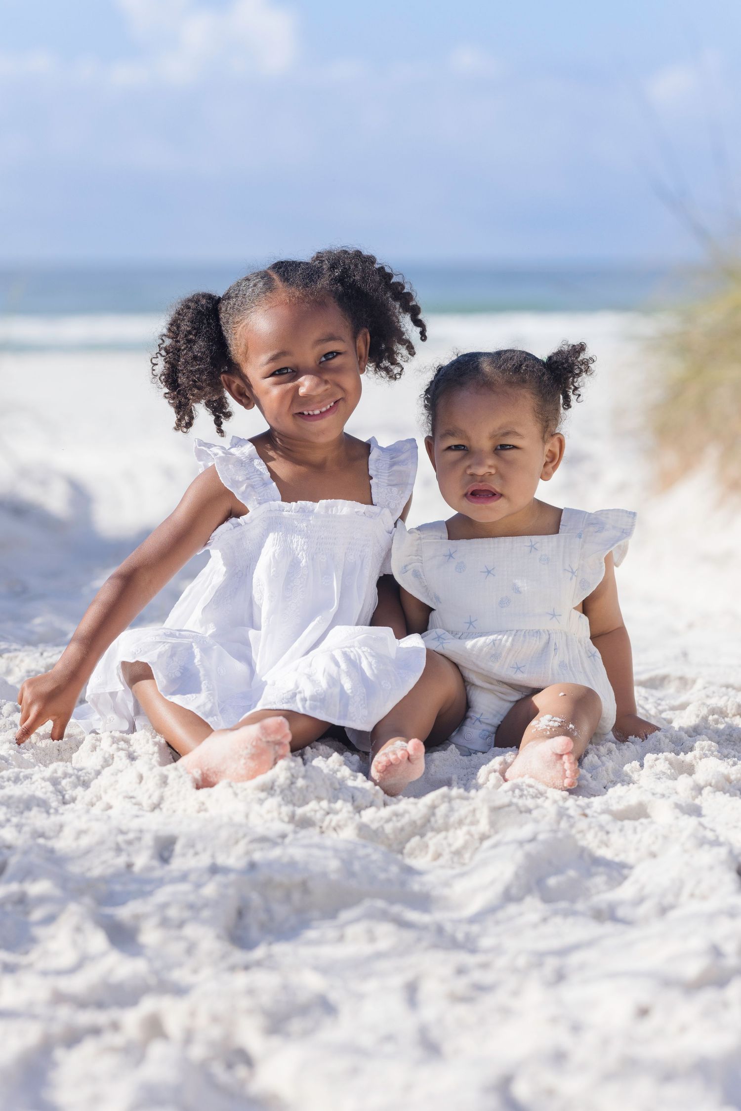 Two young African American sisters in white dresses sitting together on white sand beach, smiling on a sunny day.