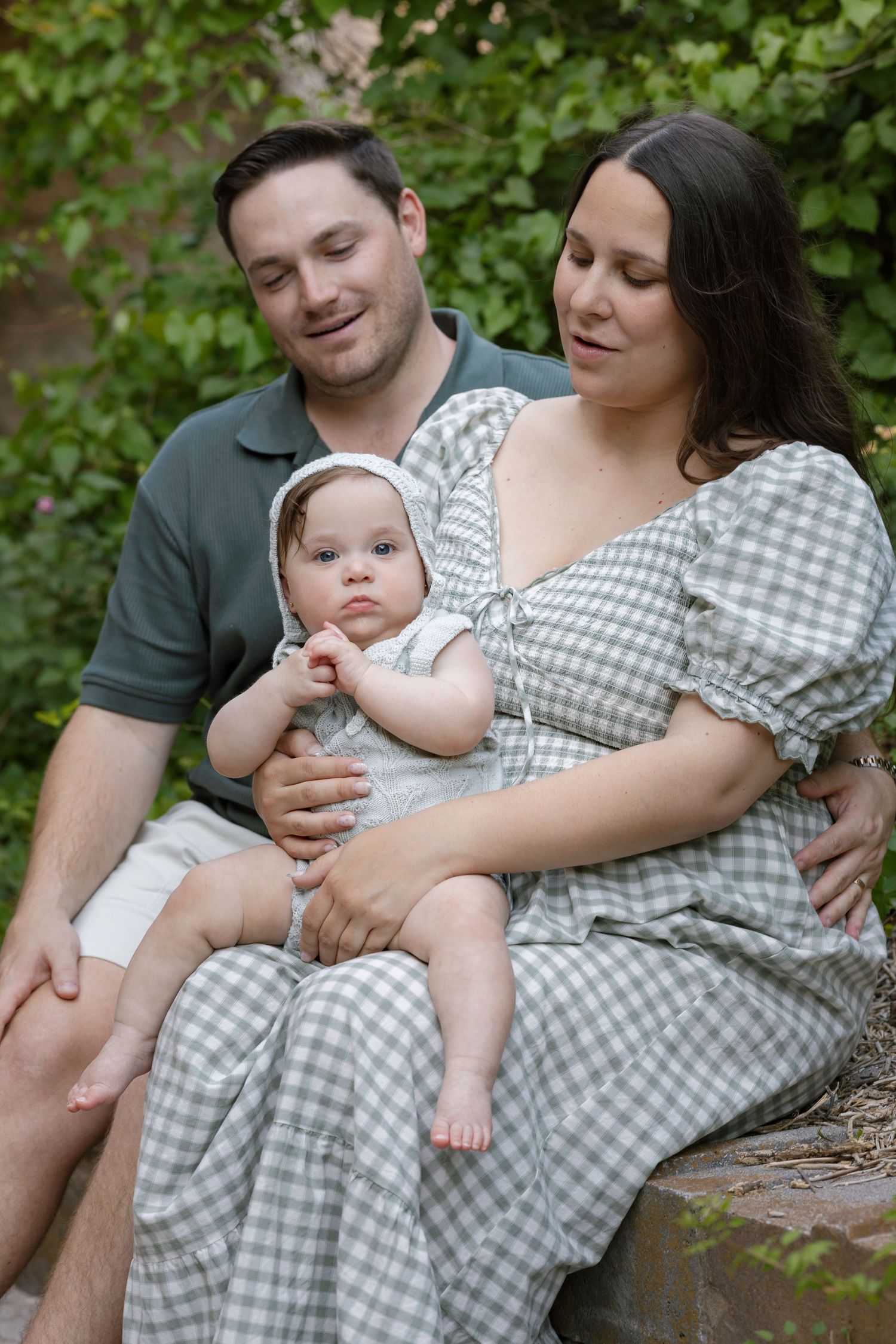 A couple sits on stone steps in a lush garden, lovingly gazing at their baby dressed in a white bonnet.