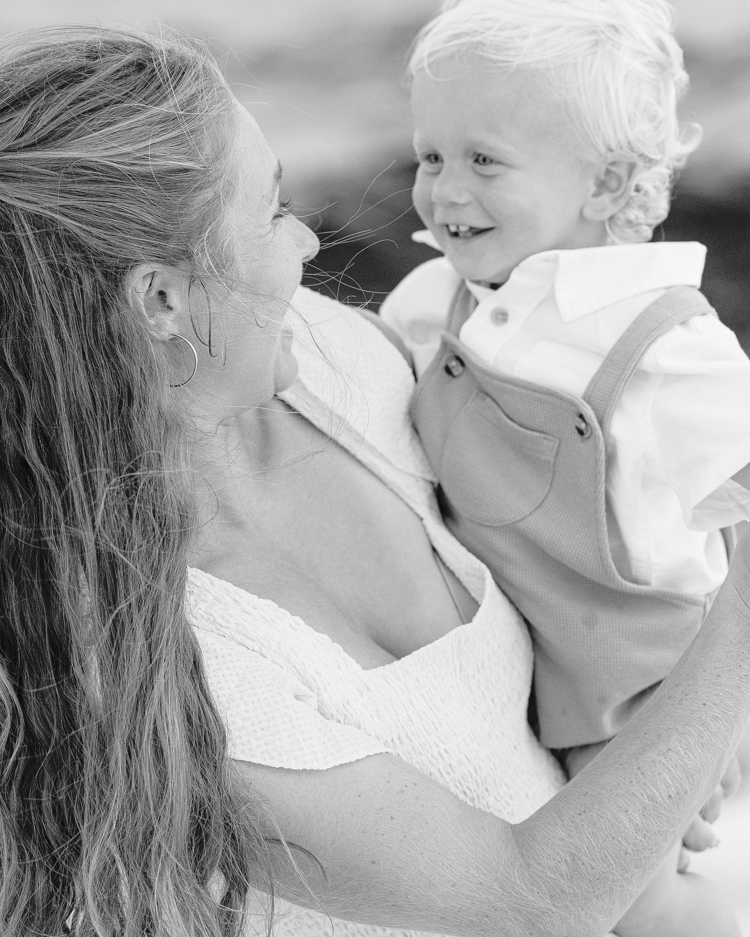Black and white photo of a smiling toddler boy in suspenders being held by a woman in a white dress.