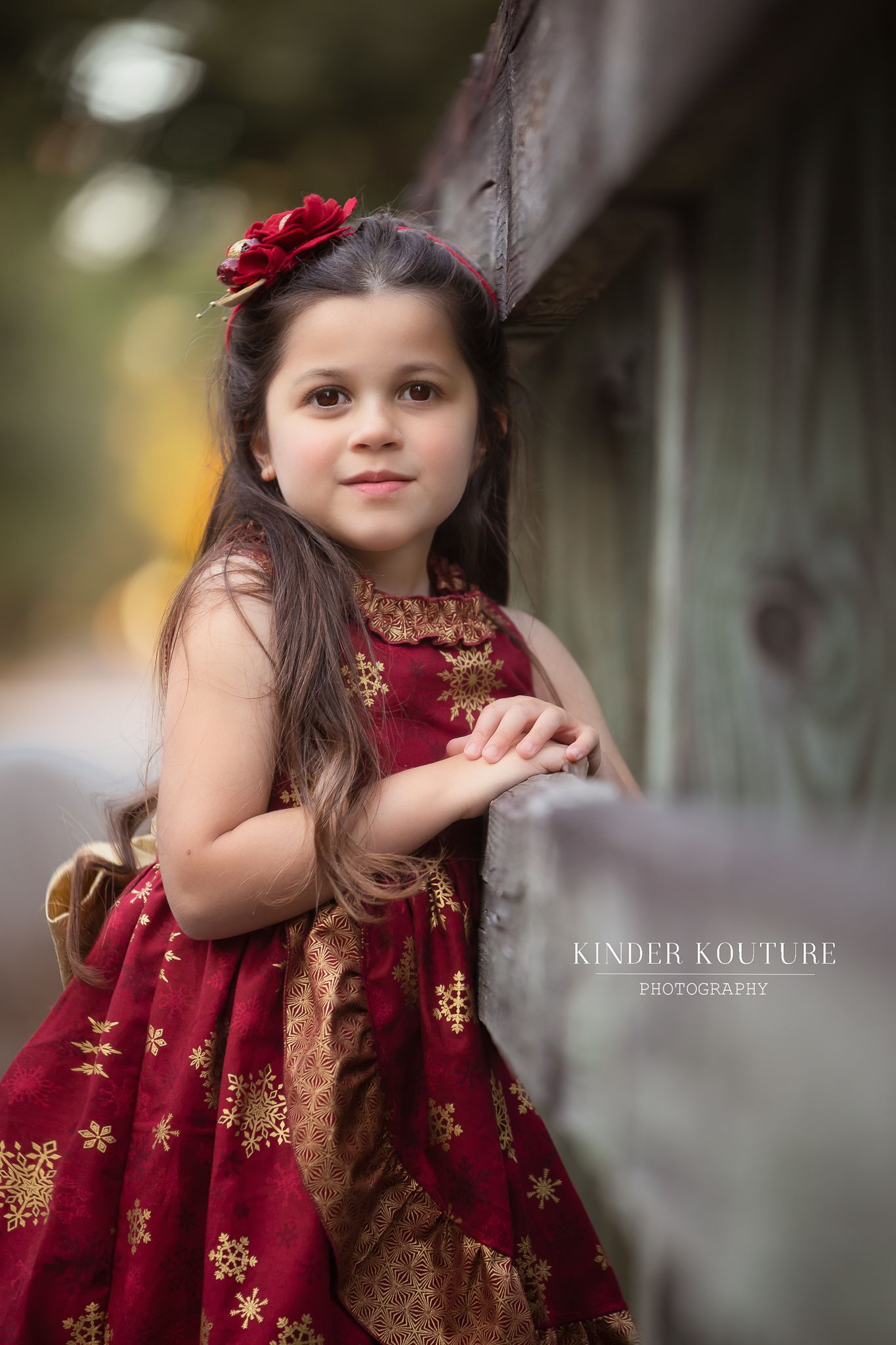 A child in an elegant burgundy and gold embroidered dress poses outdoors in a dreamy natural setting.