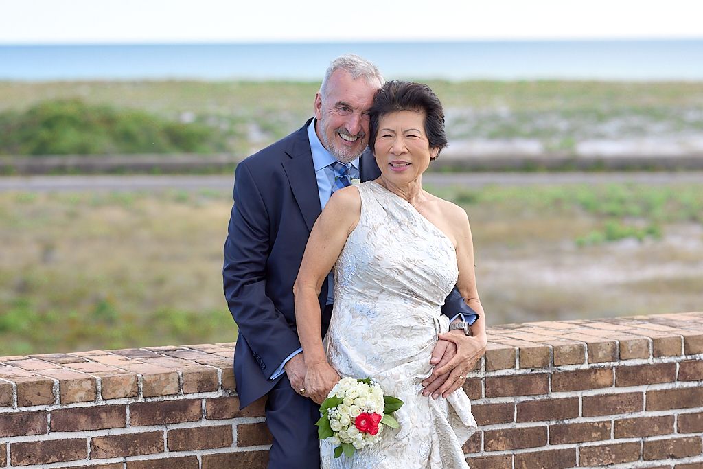 A couple in wedding attire embraces near a brick wall with a bouquet of red and white flowers.